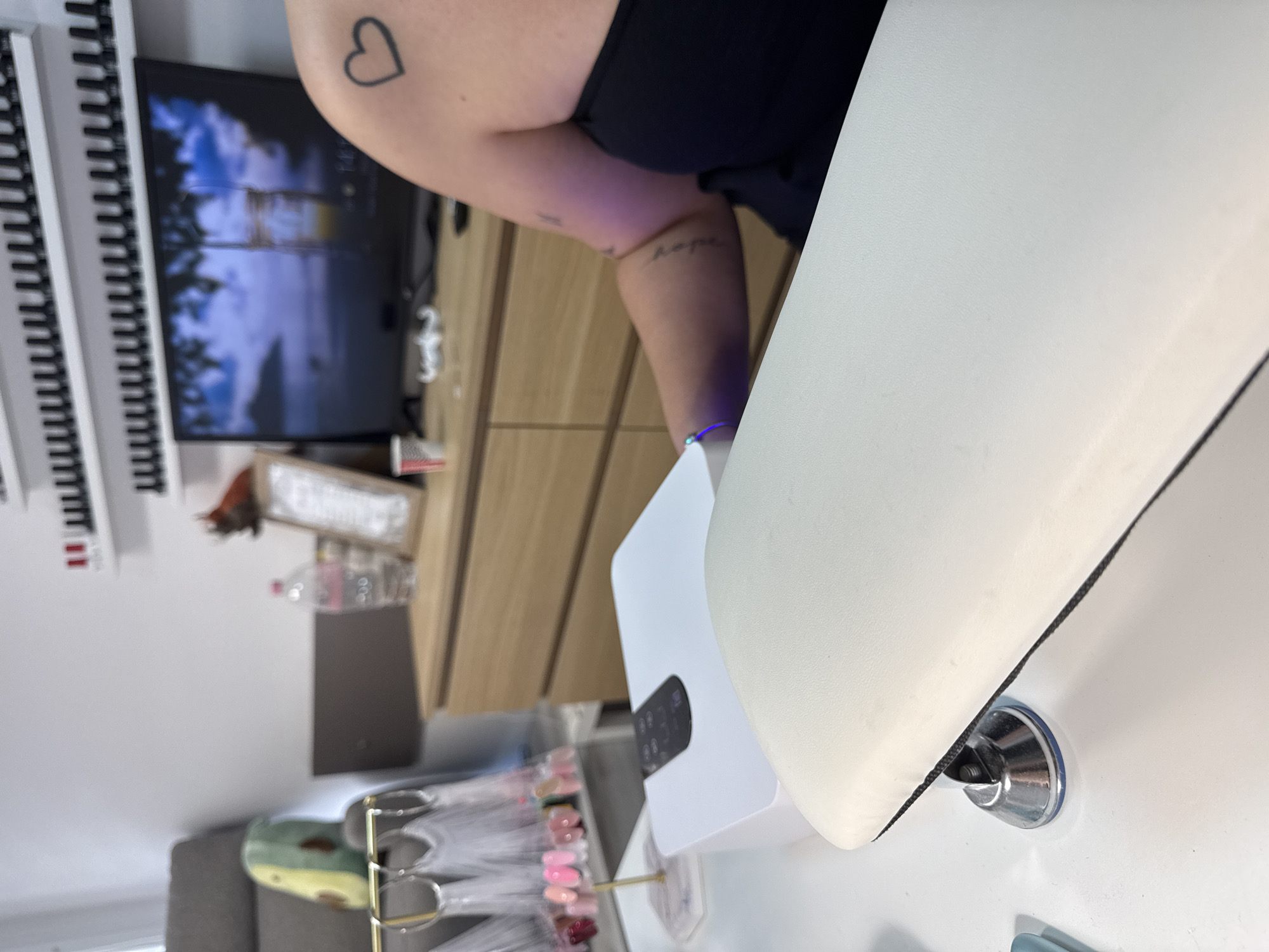 The image depicts a nail salon setting. In the foreground, a white nail lamp with a black control panel sits on a white surface. Adjacent to it is a padded white armrest. To the left, a display rack holds various nail polish swatches. In the background, a person's arm with a heart tattoo is visible, resting near the lamp. A television screen displays a tropical landscape. A wooden dresser is positioned behind the desk, and on top of it, a water bottle and a framed picture can be seen. Shelves lined with nail polish bottles are mounted on the wall. The environment appears to be an indoor commercial space, likely a nail salon. There is no information or visual cues indicating the location in Burgas, Bulgaria.