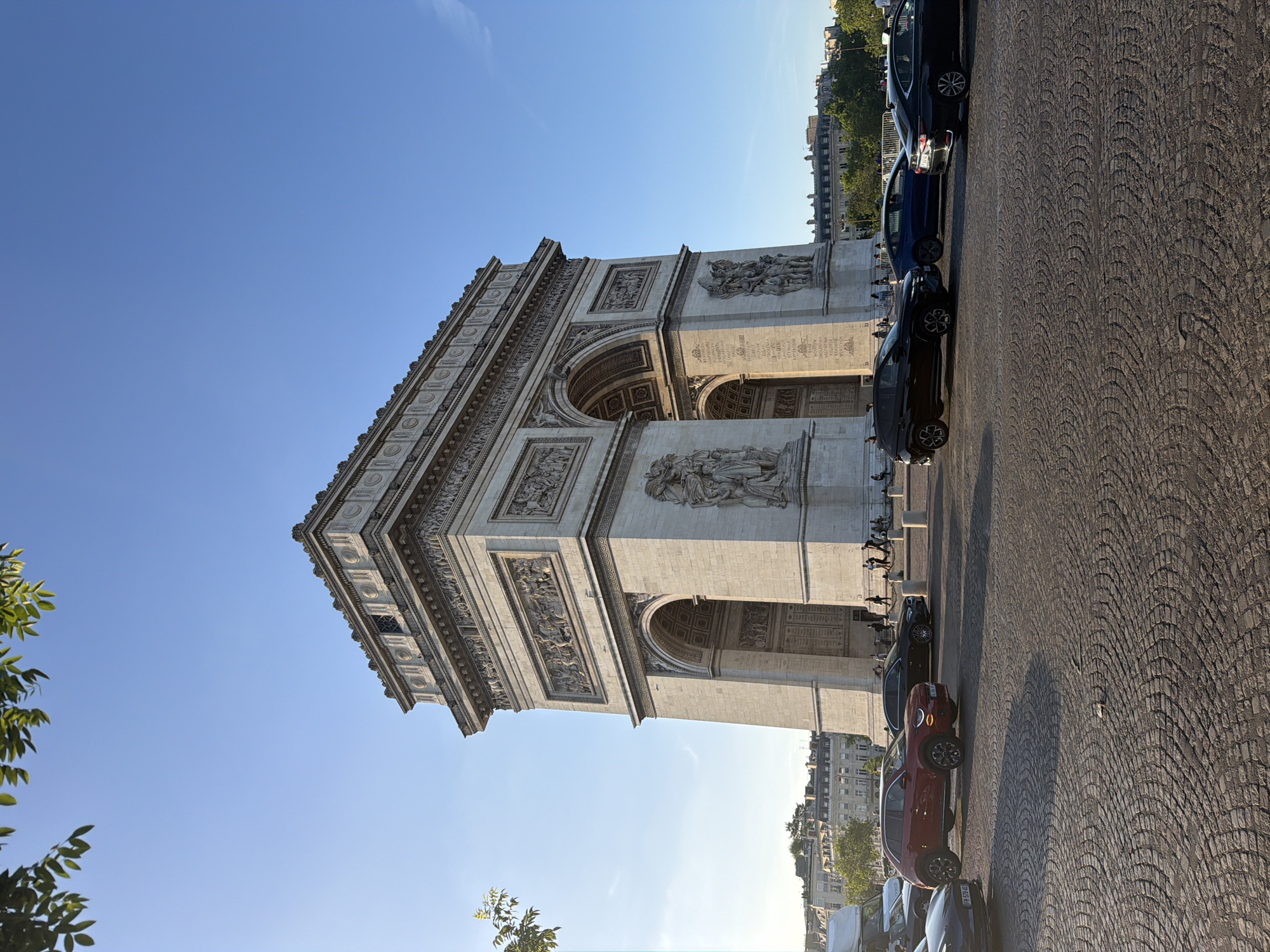 The image shows the Arc de Triomphe, a prominent monument in Paris, France. The perspective is a low-angle shot, looking up at the monument against a clear blue sky. The Arc de Triomphe is constructed of light-colored stone, adorned with detailed sculptures and architectural elements. To the right of the monument, a row of parked cars lines the cobblestone street. In the background, buildings can be seen, suggesting the urban setting of the Champs-Élysées.