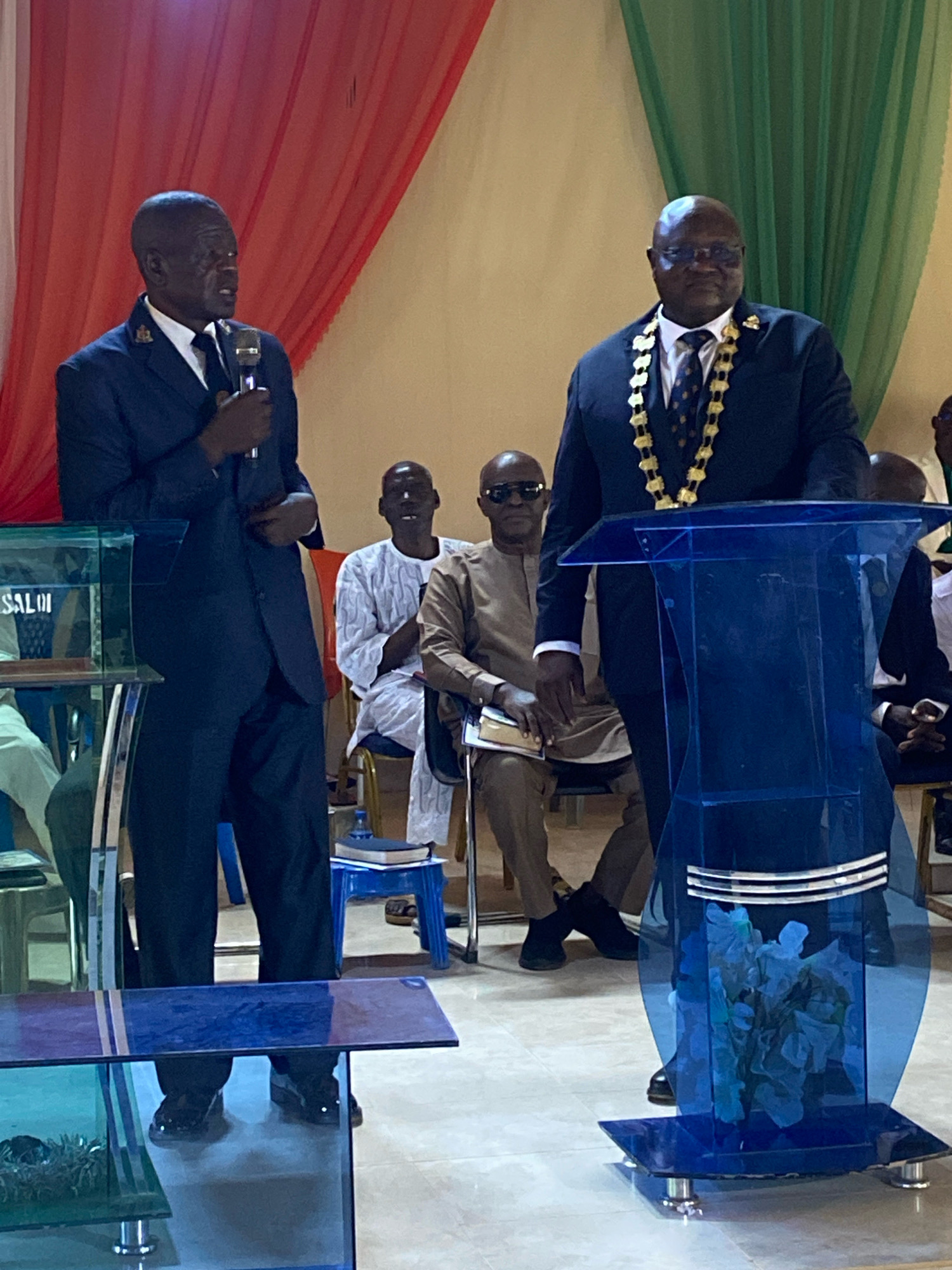 In Binchi, Nigeria, two men stand on a stage in front of a blue podium. The man on the left, wearing a dark blue suit and tie, holds a microphone and appears to be speaking. The man on the right, also in a dark suit and tie, wears a decorative gold chain around his neck and looks forward. Several other individuals are seated behind them, some in traditional attire. The backdrop consists of red and green draped fabric. The environment appears to be an indoor gathering, possibly a church or community hall, with a light-colored floor and ambient lighting.