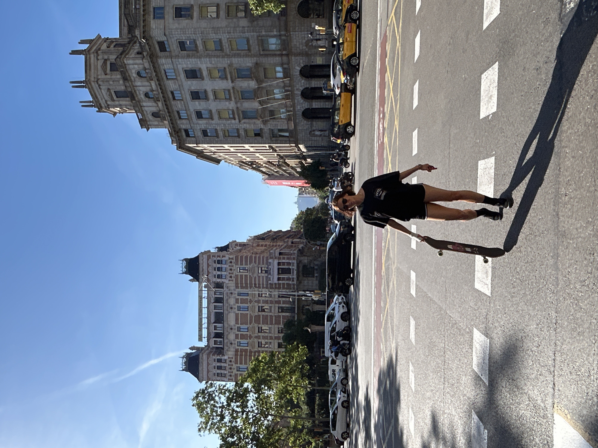 The image shows a person standing on a street in Barcelona, Spain, holding a skateboard. The person is wearing a black t-shirt, black shorts, and black boots. They are standing on a road surface with yellow and white road markings. Behind the person are buildings of varying heights, typical of Barcelona architecture. Cars and motorbikes are parked along the side of the street. The sky is blue with light cloud cover, indicating it is daytime. Shadows from trees and the person are cast on the road surface.