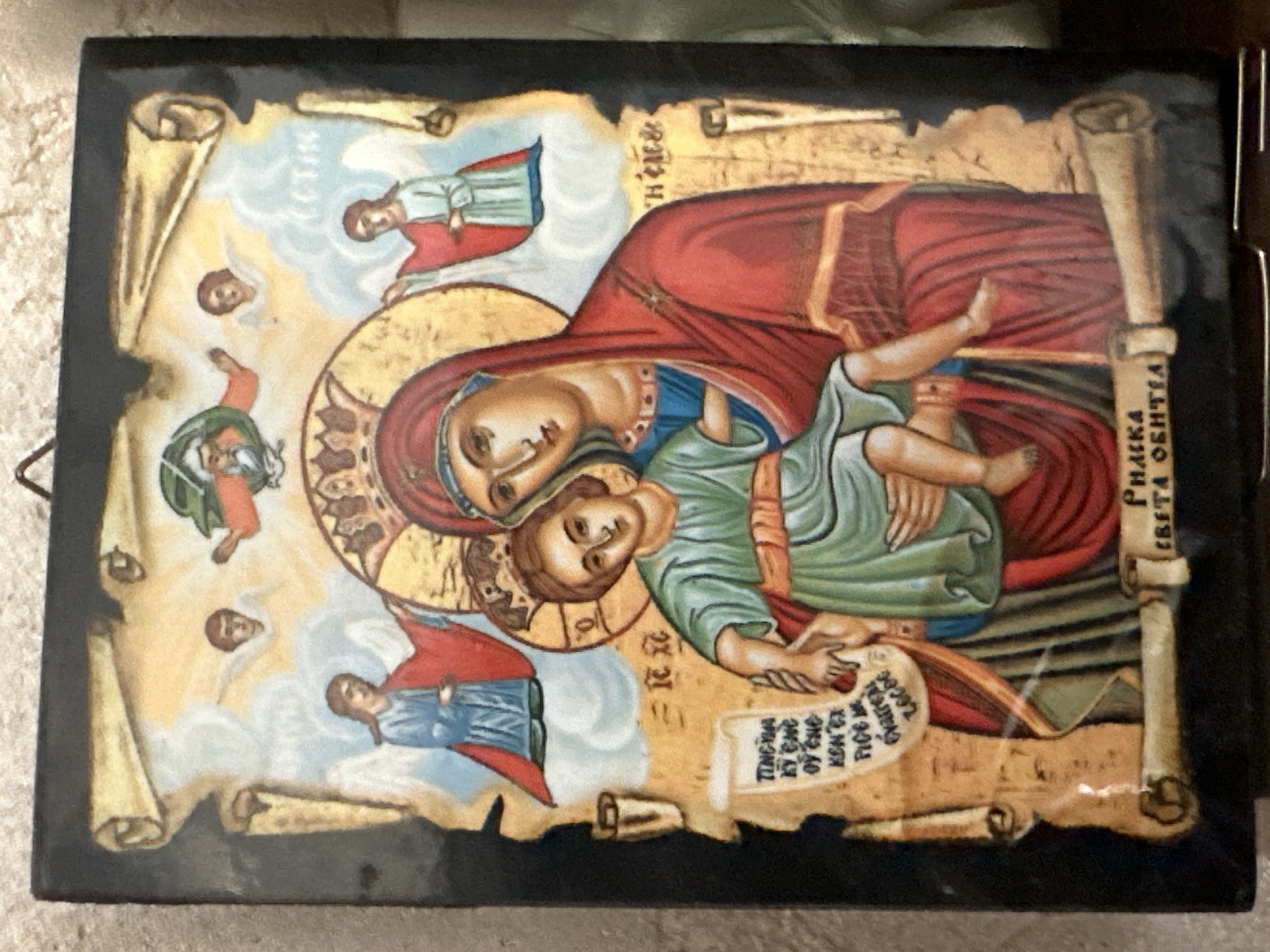 An icon depicts the Virgin Mary holding the Christ child. Both figures are depicted in a traditional Byzantine style. The Virgin Mary is wearing a red robe and a blue veil, while the Christ child is wearing a green tunic. They are surrounded by a golden halo. Above them, there are several angels. In the background, there are clouds and scrolls with inscriptions. At the bottom of the icon, there is a scroll with Cyrillic text. The icon is framed by a black border. The icon is mounted on a wooden panel and has a wire hanger. The background is a neutral, speckled surface, suggesting it is displayed indoors. The lighting is even, with no strong shadows, indicating it is illuminated by ambient light. The colors are rich and vibrant, characteristic of religious icons.