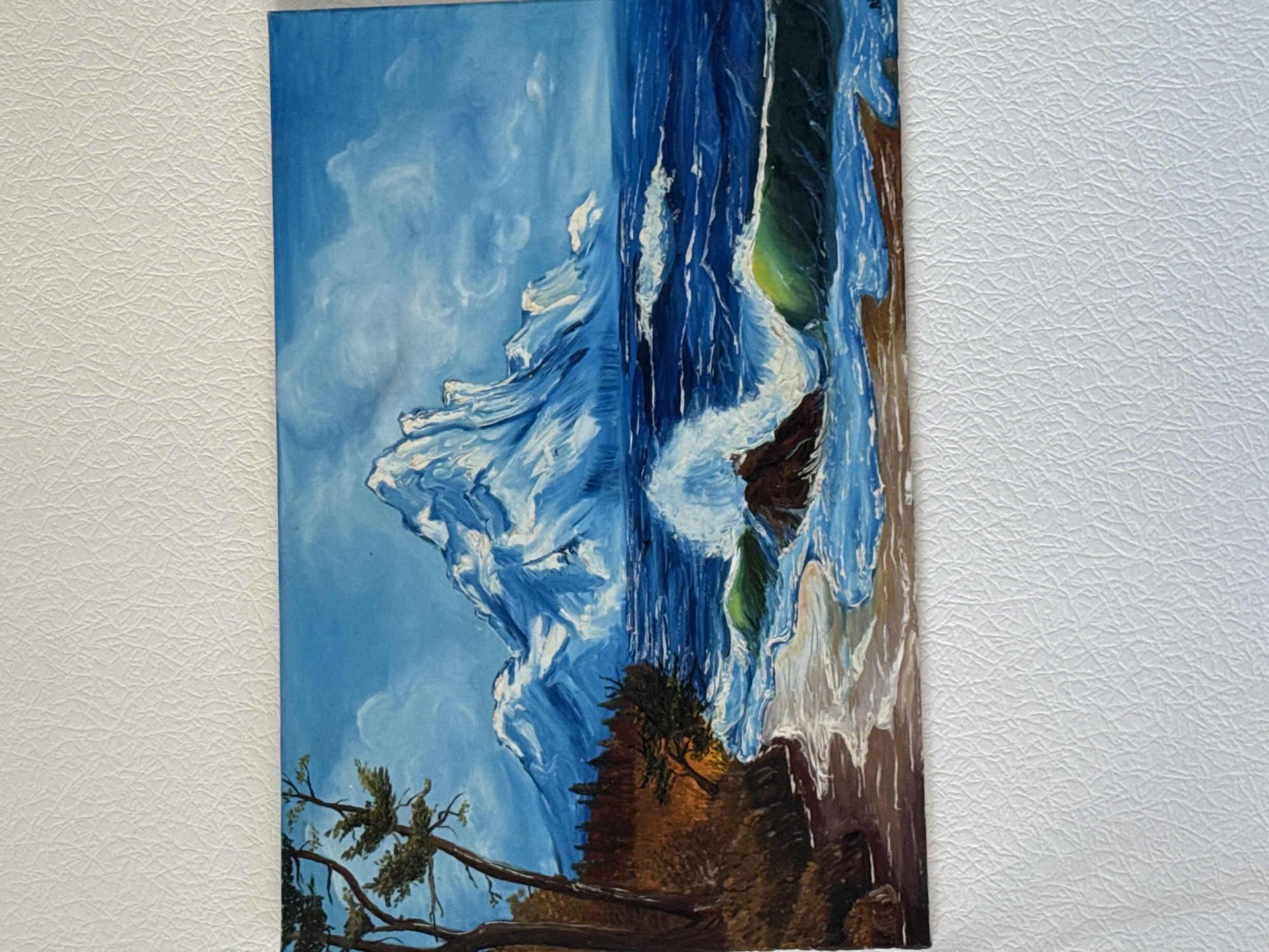 A painted landscape depicts a rugged coastline with trees and foliage on the left, leading to a body of water with crashing waves. In the background, a large snow-capped mountain range rises majestically under a cloudy blue sky. The foreground shows a sandy shore with foamy water lapping at it, and dark rocks are visible in the surf. The style is impressionistic, with visible brushstrokes and a vibrant color palette emphasizing blues, whites, and earthy browns. The painting is displayed on a white textured surface, likely a wall or a refrigerator, in an indoor setting.