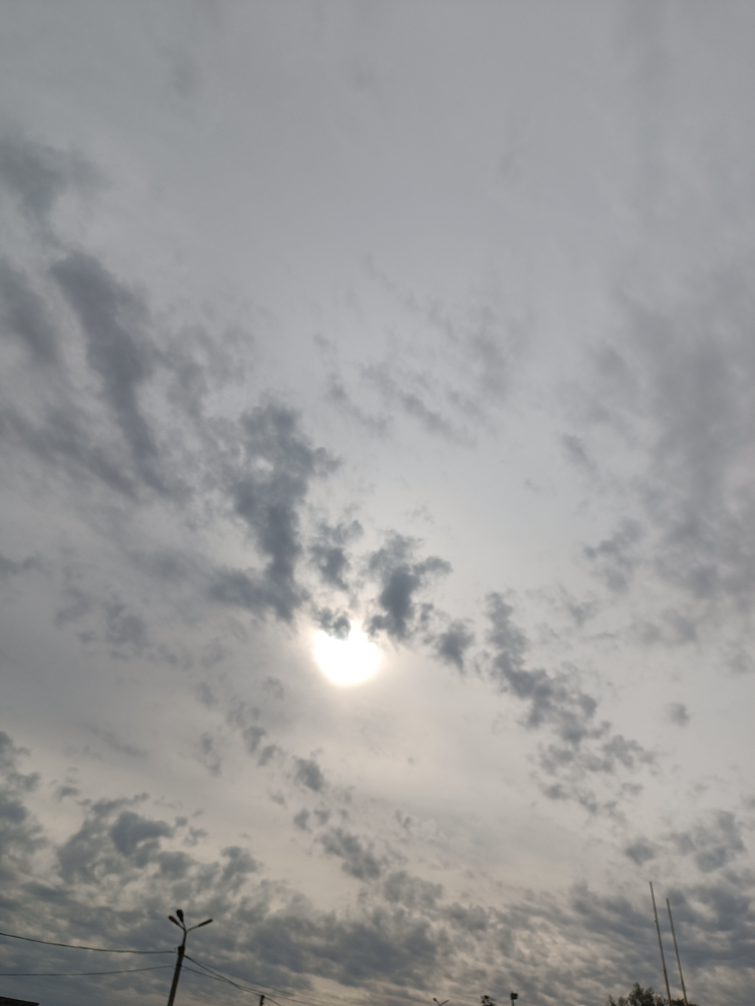 The image shows a view of the sky in what appears to be the city of Chaikovsky, Russia. The sky is predominantly gray with numerous clouds, and the sun is visible through a break in the cloud cover. In the lower portion of the image, there are streetlights and what seems to be some vegetation and a building in the distance. There are no people visible in the image. The environment seems overcast.