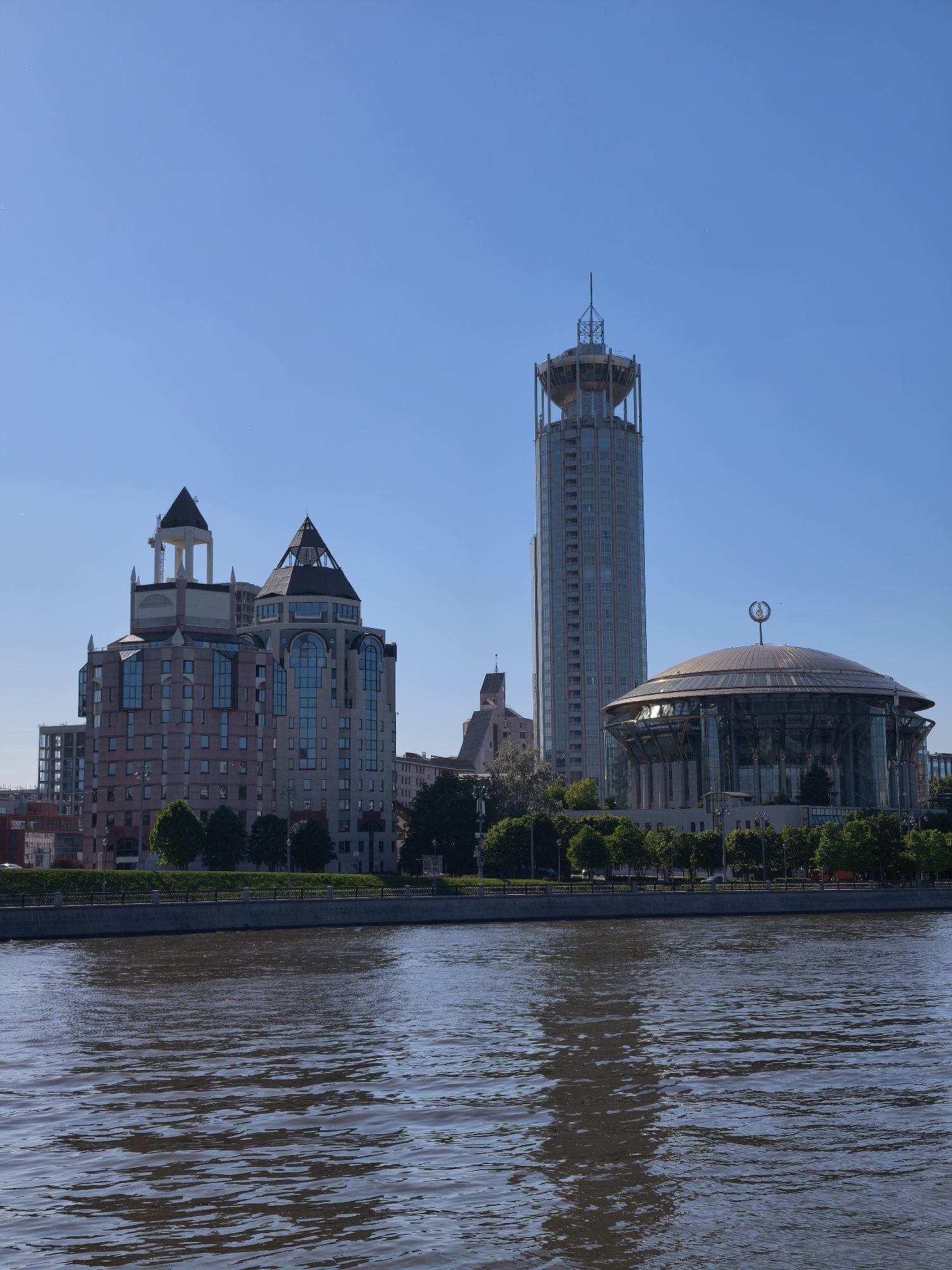 The image depicts an urban waterfront scene in Moscow, Russia, under a clear blue sky. The lower half of the frame is dominated by the Moskva River, whose dark, murky brown water exhibits numerous ripples and subtle reflections. A concrete embankment, topped with a dark metal fence or railing, separates the river from the land.
Above the embankment, a strip of vibrant green grass is visible, followed by a linear arrangement of mature, leafy deciduous trees. Behind these trees, a collection of multi-story buildings forms the cityscape.
From left to right, the prominent architectural elements include:
1. Two multi-story buildings with complex designs, featuring multiple pitched, dark grey roofs and varying facade materials, including light-colored stone and extensive glass sections. One of these buildings has a lighter, pointed structure atop its roof. These are identifiable as part of the Riverside Towers complex.
2. A tall, slender, cylindrical skyscraper, the Swissôtel Krasnye Holmy, characterized by a distinctive, open, cage-like structure at its zenith, culminating in a central spire. Its facade is primarily composed of light-colored panels and numerous windows.
3. A large, contemporary, dome-shaped building with a glass and metal exterior, the Moscow International House of Music. A distinctive circular emblem or sculpture is affixed to its apex.
The sky is uniformly clear and bright blue, devoid of clouds. Small, white, pinprick-like artifacts are present across the sky, particularly noticeable in the cropped sections, suggesting sensor dust or small specks. The scene is brightly illuminated by direct sunlight, casting some shadows. No discernible human figures are present.