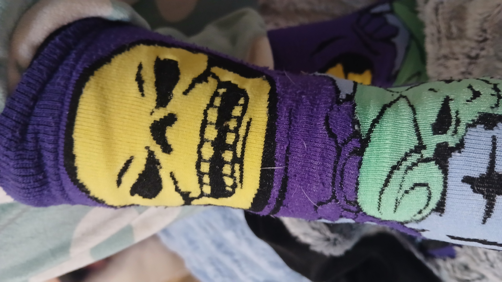 A close-up photograph shows a purple sock with a yellow Skeletor face printed on it. Another sock with a green face is also visible. The socks are crumpled in an indoor environment amongst a mix of fabrics and clothing.