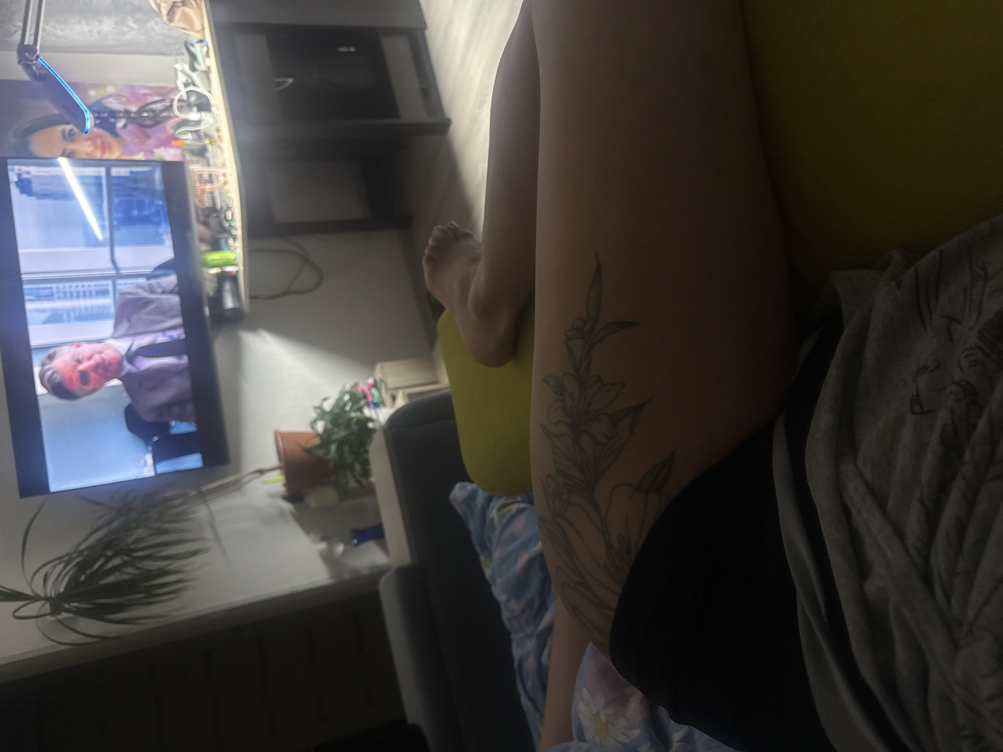 An indoor, low-angle, and tilted shot captures a person relaxing on a sofa, watching television in a dimly lit room, likely during the evening. The main subject is the person's legs and torso, seen from a personal perspective. A detailed floral tattoo is visible on the upper right thigh or hip of the person. They are dressed in a grey patterned shirt, black shorts, and partially covered by a light blue patterned blanket or sleepwear.
The television screen is displaying a scene with a man in a suit, appearing to be an anchor or interviewee, shown in the lower part of the screen. Above him, what appears to be a reflection or a secondary screen shows a woman's face, smiling. To the left of the television, a smartphone with a distinctive blue light or reflection on its edge is visible, possibly charging or actively in use. Above the TV, a shelf holds various electronic components and wires.
The room has artificial lighting, primarily from the television and possibly the phone. A potted plant is placed on a surface next to the TV, and another plant with long, thin leaves is visible on a surface in the foreground. The overall atmosphere suggests a relaxed, casual setting, possibly a living room. No specific text is clearly discernible within the image. The location is Kauchuk Stantsiya, Russia.