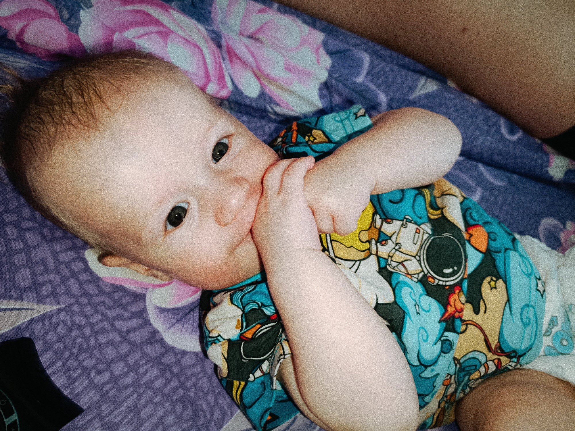 A baby is lying on a patterned purple and pink floral blanket. The baby is wearing a blue, yellow, and white shirt with a cartoon astronaut design. The baby is fair-skinned and has light brown hair. The baby is looking directly at the camera with both hands near their face, fingers possibly in their mouth. Part of an adult's bare arm is visible in the upper right corner of the frame. A black rectangular object is visible on the lower left side of the frame. The location is not provided in the image.
