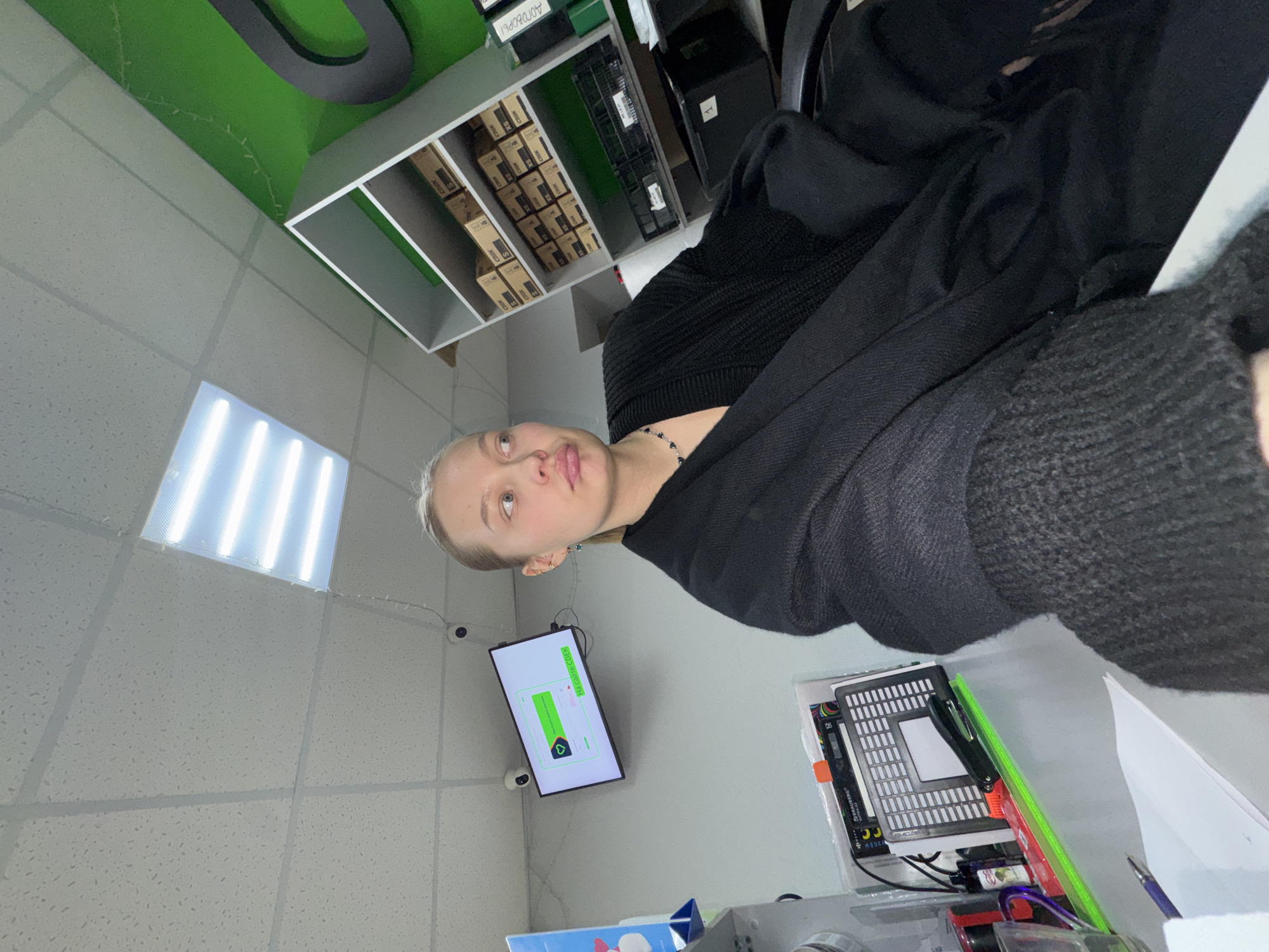 A woman is in a room with a television screen displaying "HACK CHECKER." A shelf filled with boxes is in the background. A security camera is mounted on the ceiling. The location is Chaikovsky, Russia.