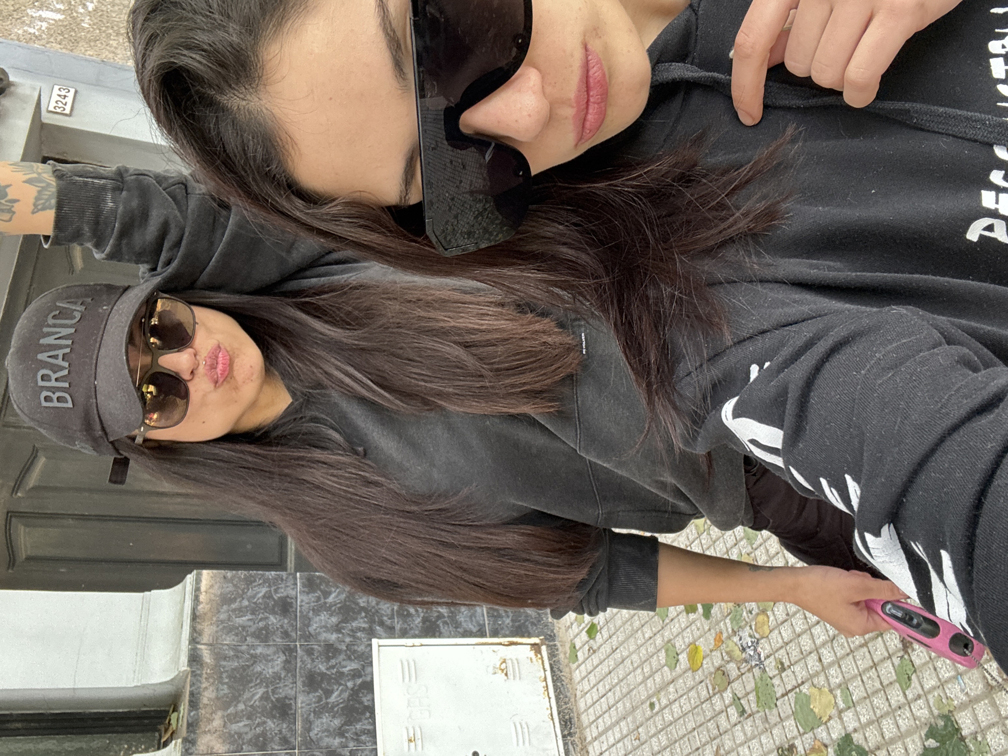 Two women are posing for a selfie outdoors. The woman in the foreground is wearing a gray baseball cap with the word "BRANCA" on it, sunglasses, and a gray hoodie. The woman in the background is wearing sunglasses and a black hoodie with white lettering on the sleeve. The location appears to be in Buenos Aires, Argentina, based on the street number "3243" visible on the wall. The ground is tiled and scattered with leaves. One of the women is holding a pink object in her hand.