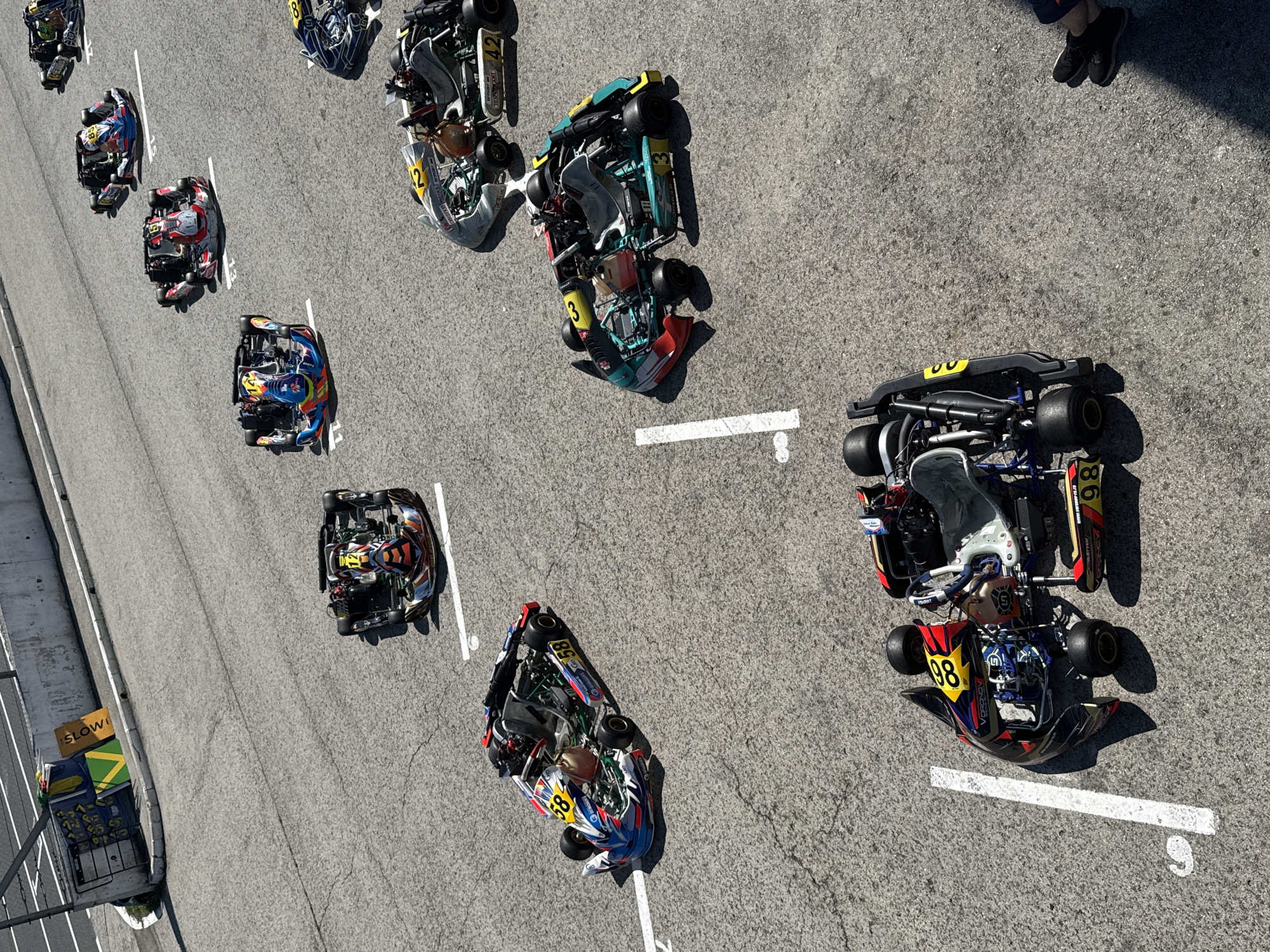 The image shows a fleet of go-karts parked on a paved surface, likely a racetrack. The go-karts are colorful and varied in design, with some featuring sponsor logos and numbers. Several white lines are painted on the ground, indicating a track or parking stalls. In the background, there is a concrete barrier and a sign with the word "SLOW" visible. The overall lighting suggests a sunny day. The location is identified as Sekiotovo, Russia. There are no people actively driving or interacting with the go-karts in this specific frame, though the presence of a person's feet in the lower right corner suggests at least one individual is present nearby.