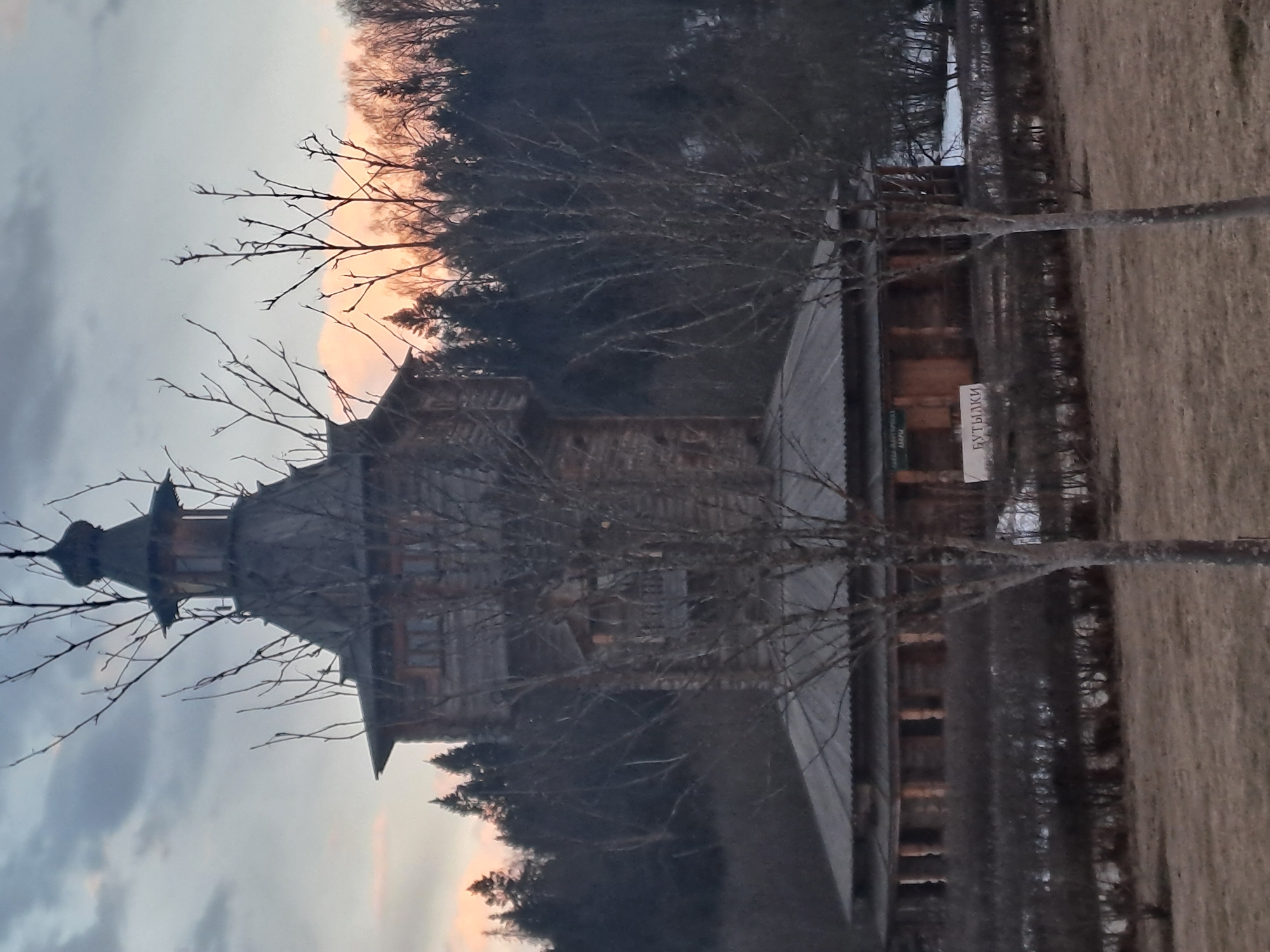 The image shows a tall, wooden building with a spire, possibly a church, in Vzglyadnevo, Russia. Bare trees obscure parts of the structure. A sign with Cyrillic text is visible near the building's entrance. The environment includes a grassy field and a dark forest in the background. The sky is a mix of grey and orange, suggesting either sunrise or sunset. No people are visible.