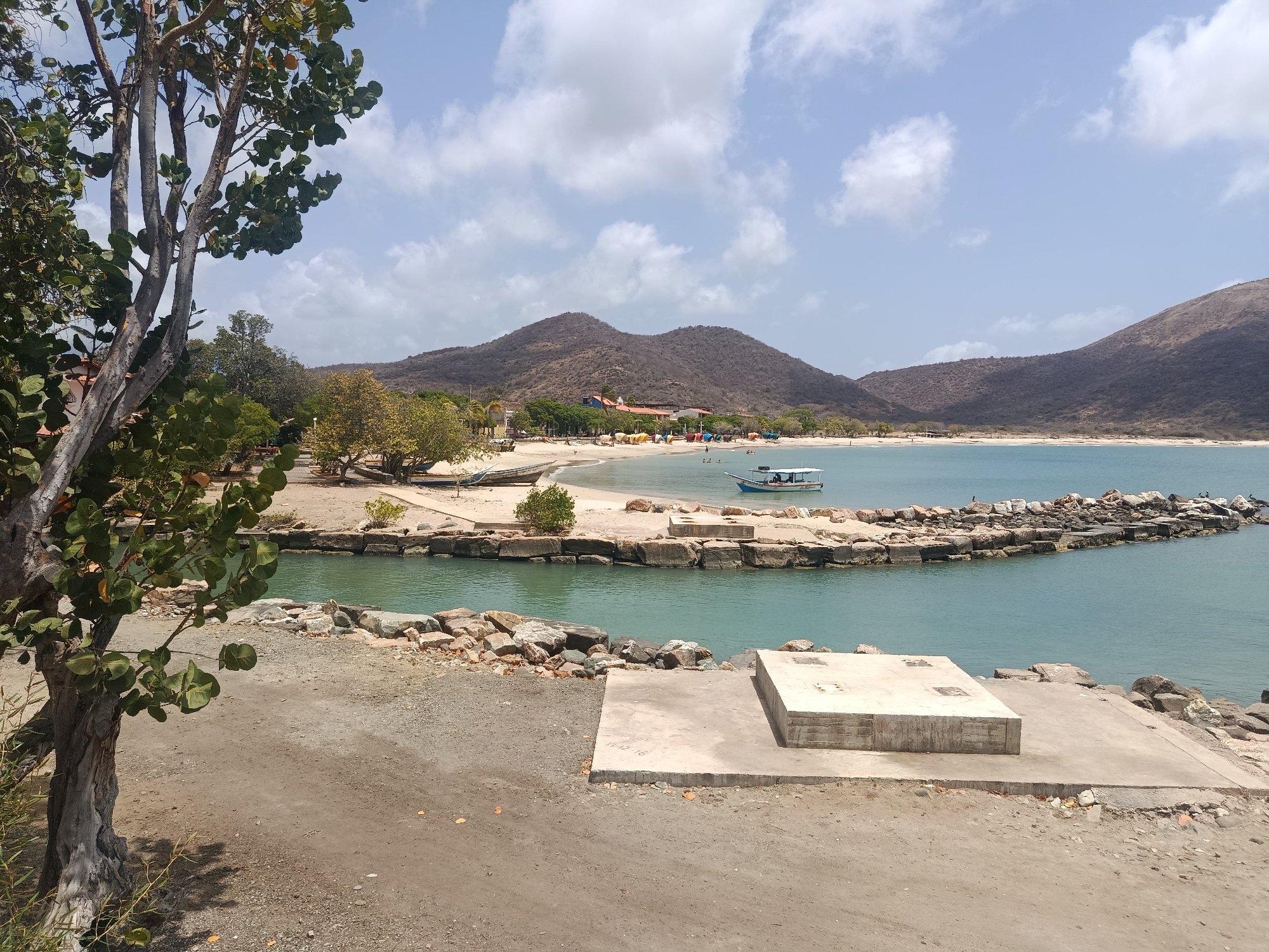 The image depicts a coastal scene in Pedro González, Venezuela. In the foreground, there's a sandy area with a concrete platform and block. A partially visible tree with green leaves is on the left. Beyond the foreground, a lagoon or inlet is visible with a breakwater made of rocks extending into the water. A small boat is floating in the lagoon. A sandy beach stretches across the middle ground, with people visible on the beach. In the background, hills rise against a partly cloudy sky.