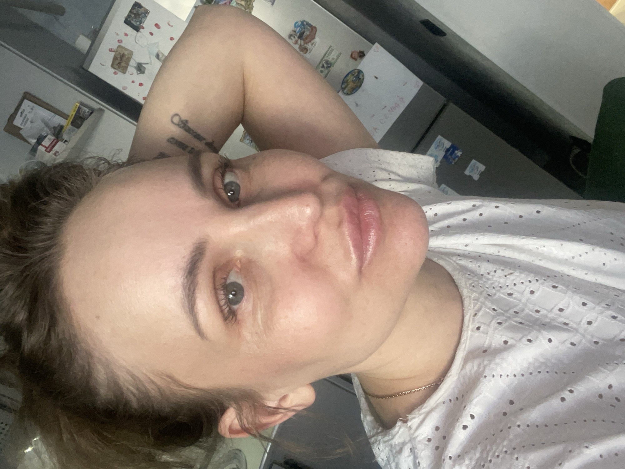 A woman is posing for a selfie indoors. She is wearing a white eyelet top and a gold chain necklace. Her right arm is raised above her head, revealing a tattoo on her inner arm. She has light skin, light-colored eyes, and brown hair pulled back. The background includes a refrigerator with various magnets and papers attached. The location appears to be an interior space in the city of Chaikovsky, Russia.
