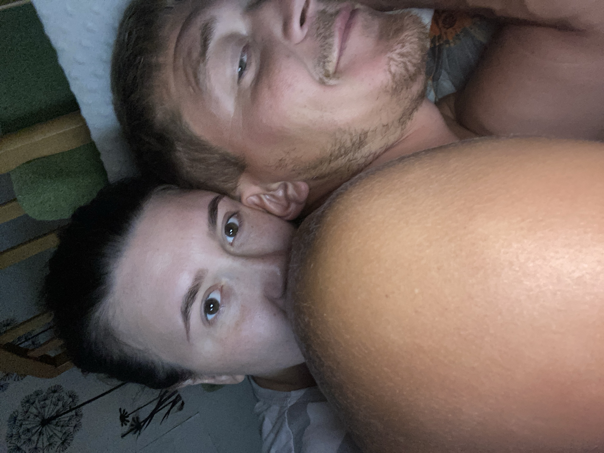 The image shows two people, a woman and a man, in close proximity, likely in bed. The woman's face is partially visible, with her eyes looking towards the camera. The man's face is also visible, with his head turned and a slight smile. They are lying down, with a large, indistinct object in the foreground, which could be an arm or a shoulder. The background appears to be a bedroom, with what looks like bedding and a wall with a pattern that resembles dandelions. There is no identifiable signage or landmarks to indicate the specific location within the city of Chaikovsky, Russia. The overall lighting is dim, suggesting it might be nighttime or the room is dark.