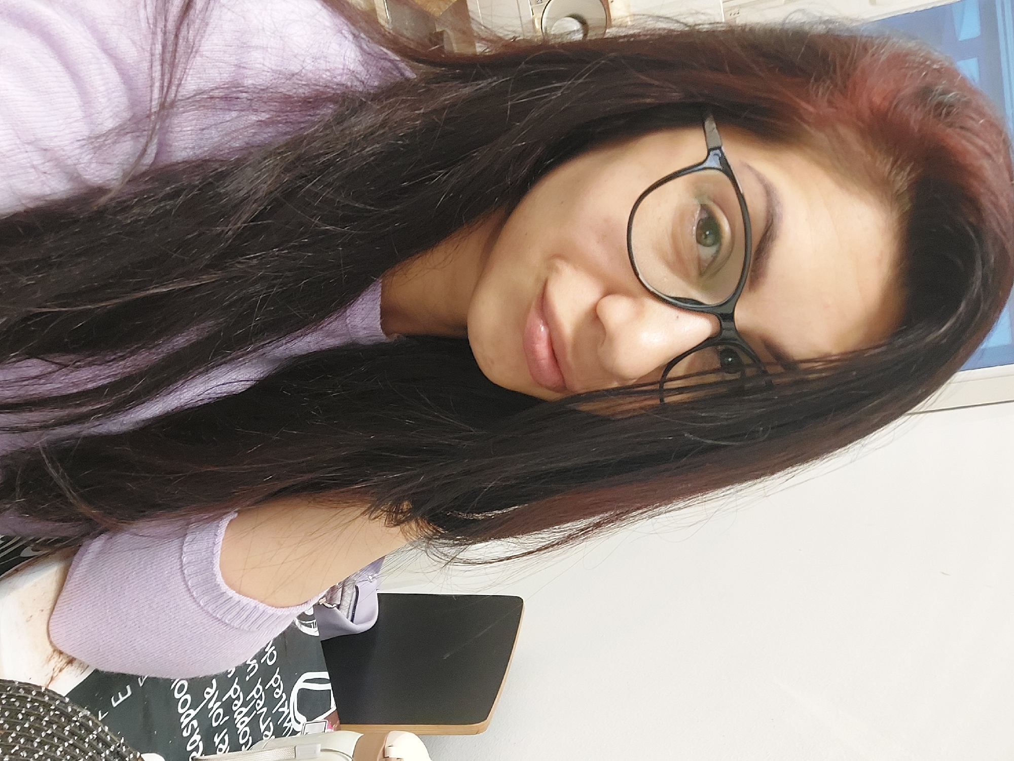 The image shows a close-up, self-taken photograph of a woman with long, dark brown hair with reddish highlights, wearing black-rimmed glasses and a light purple long-sleeved shirt. She is looking towards the camera with a slight smile. The background is a plain white wall, with some blurry elements suggesting an indoor setting. There is a black object with a wooden edge on the right side of the image, and a surface with white text on it in the lower right corner. The image does not provide any specific visual cues that would indicate its location within the city of Landshut, Germany.