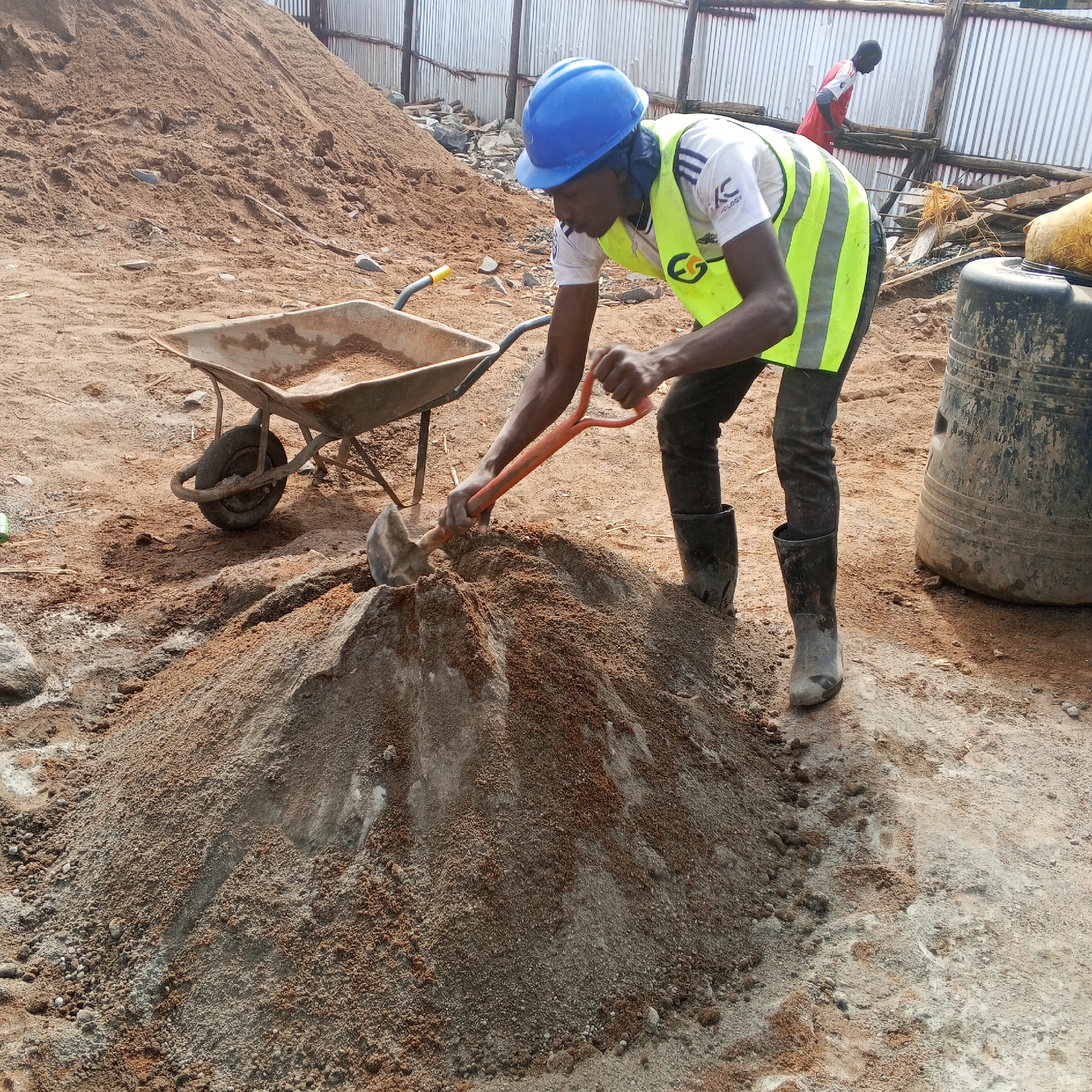 In Borabu Chitago ward, Kenya, an outdoor scene depicts a construction worker engaged in manual labor. The main subject, a man, is seen actively shoveling and mixing a large pile of sandy, earthy material on the ground. He wears a bright blue hard hat, a white t-shirt with "IC TOLOGY" printed on it, and a fluorescent yellow high-visibility vest featuring an "EG" logo. His attire includes dark trousers and black rubber boots. He uses an orange-handled shovel to work with the material.
To the left of the worker, a metal wheelbarrow rests on the dusty ground, partially filled. In the background, a massive mound of similar sandy material stretches across the left half of the image. On the right, a large, dark-colored plastic container or barrel is visible. Further back, another individual, dressed in a red shirt, is partially obscured, appearing to work near a temporary structure constructed from corrugated metal sheets and wooden planks.
The setting is a rugged construction or material handling area, indicated by the piles of raw materials and rudimentary structures. The time of day is clearly daylight, with bright, natural illumination. The ground is unpaved and uneven, covered in dust and loose earth, consistent with a busy worksite. No strong emotions are discernible from the workers; they appear focused on their tasks.