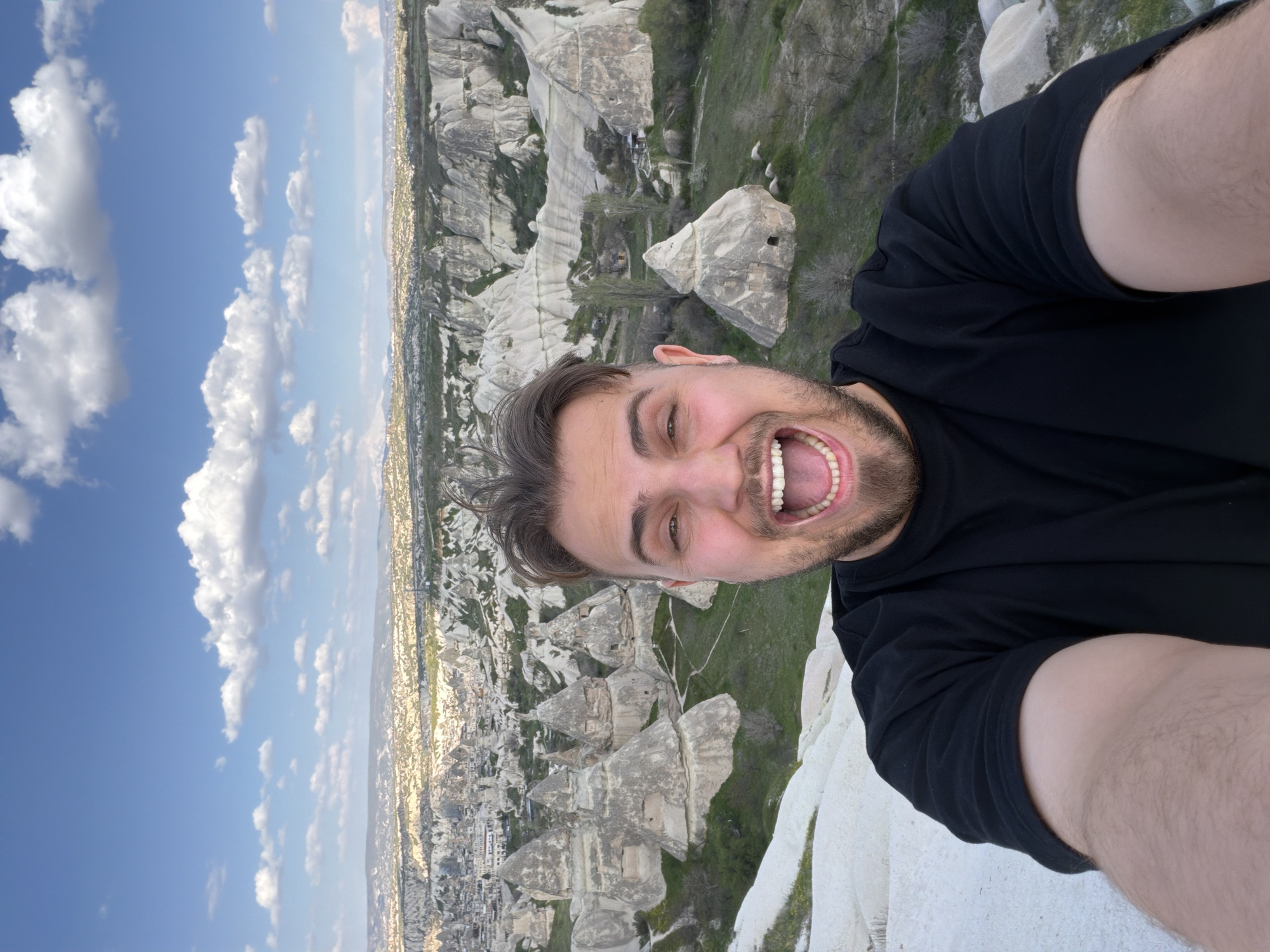 A young man is captured in a joyous selfie, positioned to overlook the unique geological landscape of Göreme, Cappadocia, Turkey. He is seen from the chest up, with his face prominent in the upper left of the frame. He appears to be in his 20s or early 30s, with dark hair and a beard, and is wearing a black short-sleeved t-shirt. His expression is one of pure delight, with a wide, open-mouthed smile revealing his teeth, and bright, happy eyes, conveying excitement or awe at his surroundings.
The background reveals the iconic "fairy chimney" rock formations and deep valleys characteristic of Göreme. These distinct, light-colored volcanic tuff structures are interspersed with areas of green vegetation. In the distance, a town or city is visible, nestled among the rock formations. The sky above is a clear, vibrant blue, dotted with scattered white, fluffy clouds, indicating bright daytime conditions, likely late morning or early afternoon. No visible text is present in the image.