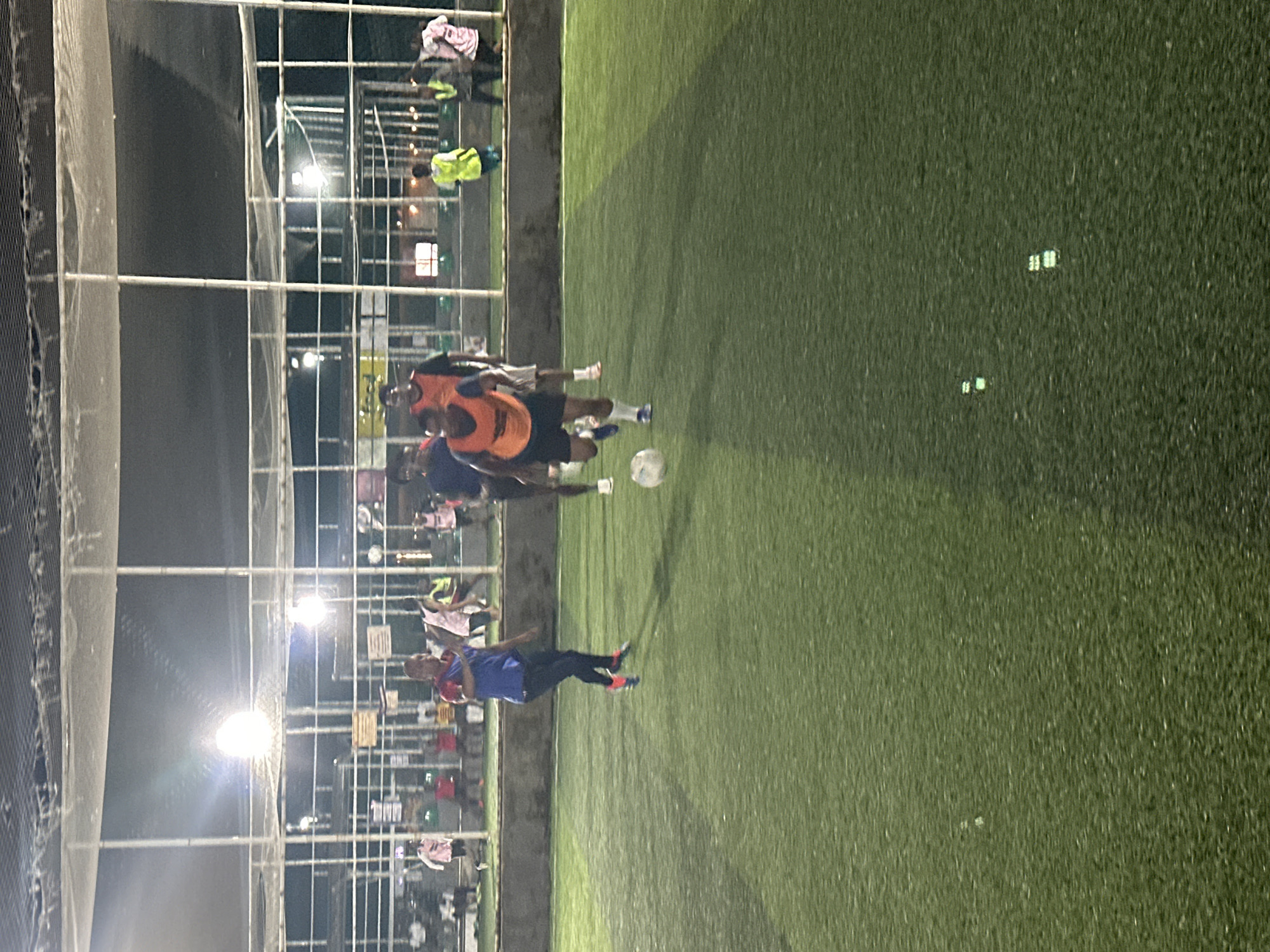 The image shows a group of people playing soccer on an artificial turf field at night. The field appears to be enclosed by netting and illuminated by overhead lights. The players are wearing various colored athletic attire, including some individuals in orange and blue vests. One player in the foreground is dribbling a soccer ball. In the background, there are spectators and other individuals standing around the perimeter of the field. The overall setting suggests an amateur or recreational soccer match taking place in an urban environment. The image is of low resolution and has a grainy quality, typical of nighttime photography. The scene is likely located in Abuja, Nigeria, as requested.