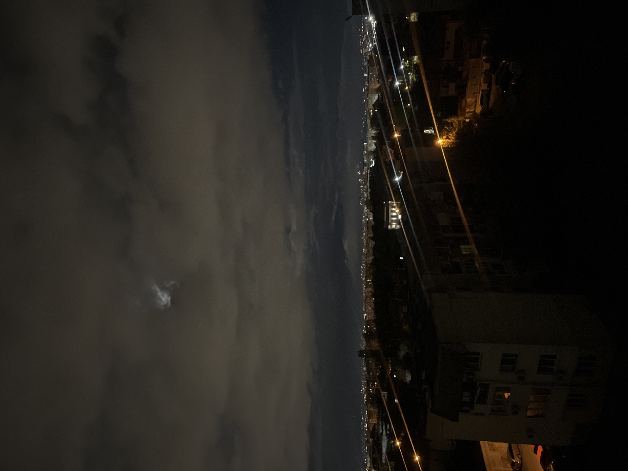 At night, the image shows a view from a high elevation, likely a building, in Sofia, Bulgaria.
To the left, clouds fill the sky with a small moon visible.
To the right, a city skyline is visible with many yellow streetlights, and a tall building is on the lower right corner.