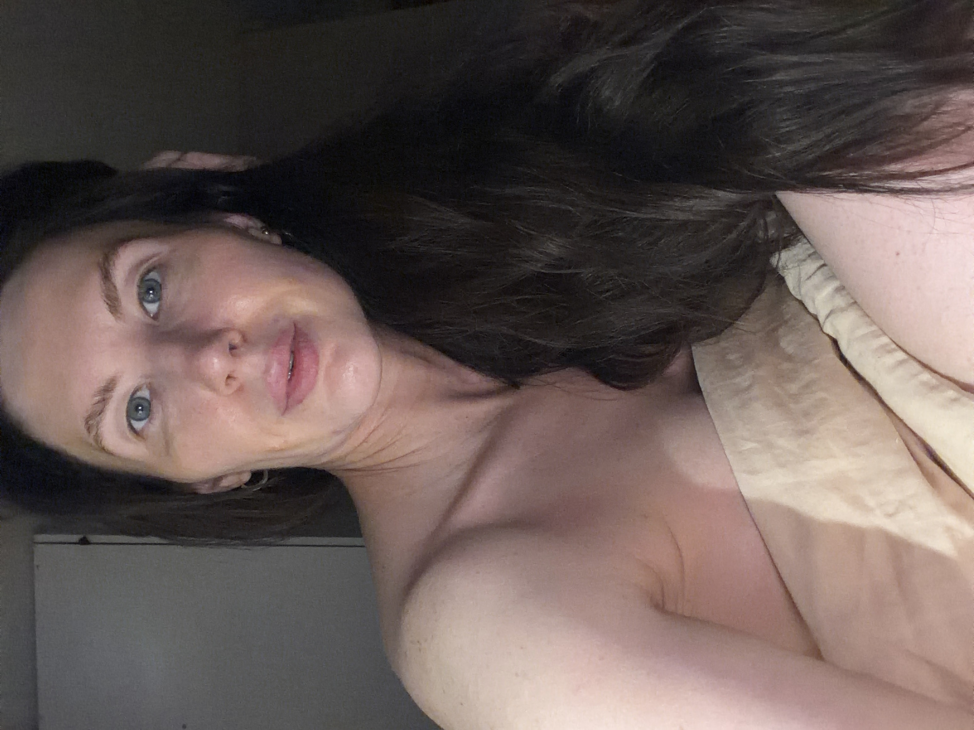 A woman with long dark hair and fair skin is reclining on a beige-colored fabric, possibly a bedsheet or blanket. Her face is turned towards the camera, revealing blue eyes and visible pores. She appears to be indoors, and the lighting is dim, with a gray wall visible in the background. There is no indication of the location being Perm, Russia, or any other specific location. The scene is focused on the woman's face and upper body, with the rest of the environment obscured by darkness.