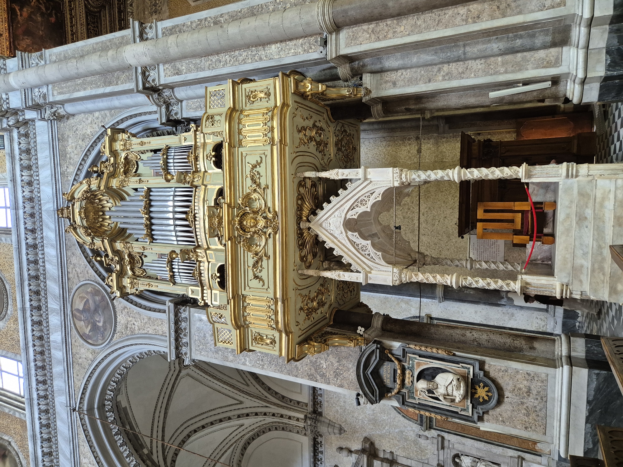The image shows an interior view of the Basilica di San Lorenzo Maggiore in Naples, Italy. In the center of the frame is a large, ornate pipe organ mounted on a decorated balcony. The organ is a light green color with extensive gold detailing and numerous silver pipes. Below the organ, there is a white, Gothic-style structure with intricate carvings and twisted columns, leading up to a raised platform. To the left of this structure, a framed relief sculpture of a robed figure holding a staff is mounted on the wall. The walls of the basilica are adorned with decorative patterns and marble-like finishes. Tall, fluted columns rise to support the vaulted ceiling, which is partially visible at the top of the frame. The floor in front of the organ structure is a black and white checkered tile pattern. A person, wearing light-colored trousers and a dark shirt, is visible standing near the right side of the white Gothic structure, partially obscured. There are wooden benches visible in the foreground. The overall scene depicts a grand and historic religious interior.