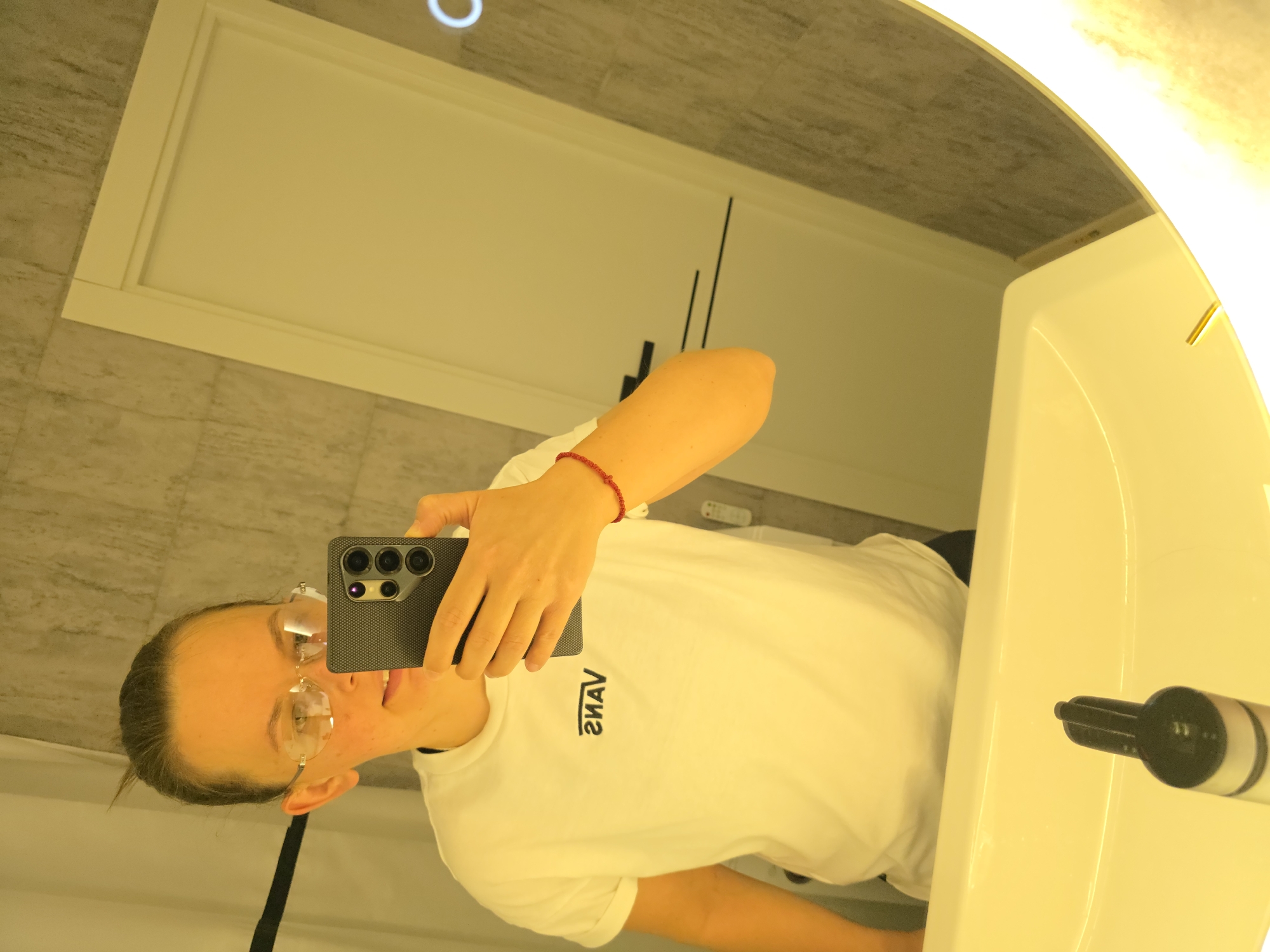 A woman wearing a white t-shirt and glasses is taking a selfie in a bathroom. She is holding a smartphone in her right hand, which is in focus. The reflection in the mirror shows her face and the upper torso of her body. She has a red bracelet on her wrist. The bathroom appears to be modern, with a tiled wall and a large illuminated mirror above a sink. The sink itself is visible in the foreground, with a dispenser on it. The image was taken in Rostov-on-Don, Russia.