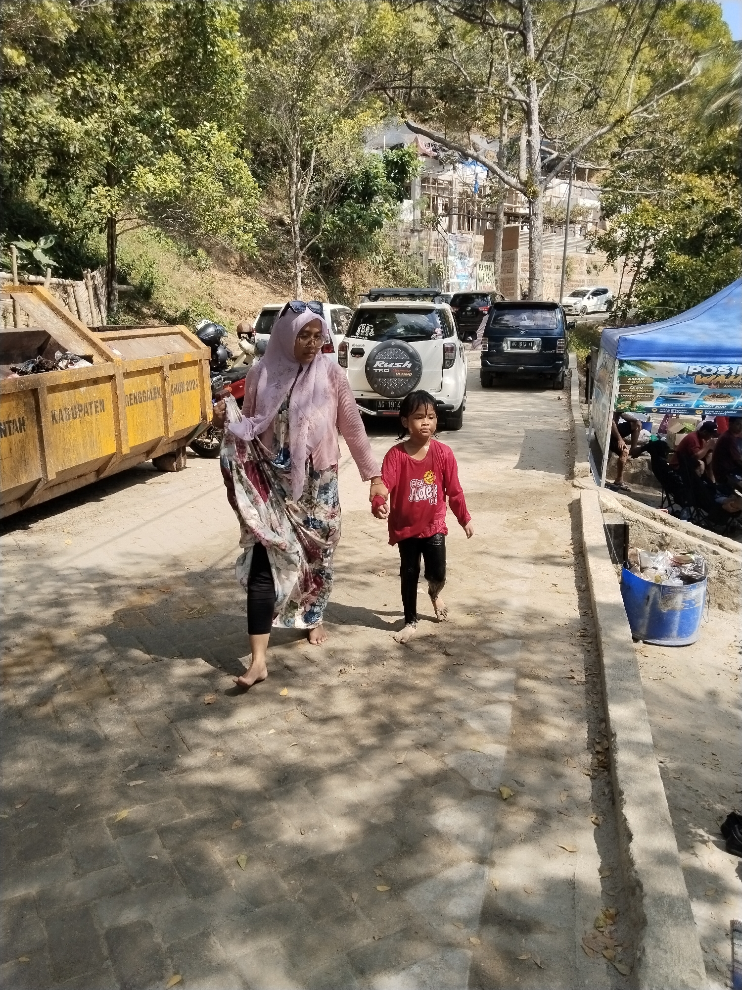 In Prigi, Indonesia, a woman wearing a pink hijab and a floral dress walks barefoot, carrying a folded cloth. Beside her, a young girl in a red long-sleeved shirt and black leggings also walks barefoot. They are on a paved walkway with shadows from trees overhead. To their left, a large yellow dumpster is visible, labeled with "KABUPATEN" and "TRENGGALEK TAHUN 2024". Behind them, several cars are parked, including white and dark-colored SUVs. To the right, a blue tent is set up, with people visible underneath and behind it, some sitting and others standing. A blue trash bin is positioned next to the tent. The ground is a mix of sand and paved surfaces, with scattered leaves. In the background, a hillside covered in trees and some structures can be seen.