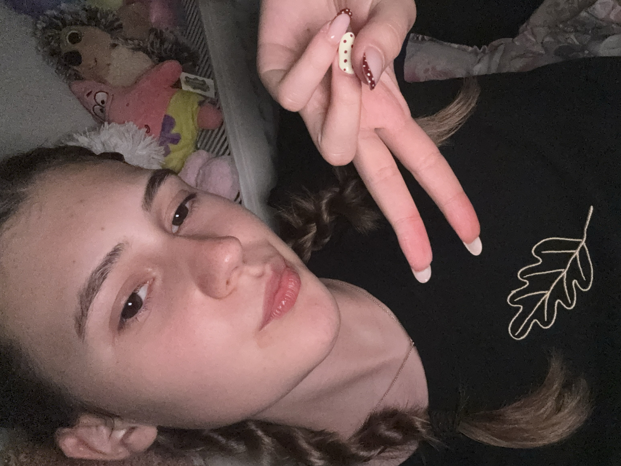 A young female, likely a teenager or young adult, is the central subject of this close-up portrait, appearing to be lying down for a selfie or photograph. Her head is tilted, with her gaze directed towards the camera. She has fair skin, dark eyes, and well-defined eyebrows. Her lips are slightly parted, and her dark brown hair is styled into two visible braids.
She wears a dark-colored top or sweatshirt, which is adorned with a light-colored embroidered outline of an oak leaf on the right side of her chest. A delicate silver chain necklace is visible around her neck. Her left hand is raised, performing a peace sign gesture. Her manicured fingernails display distinct designs: one nail is light with small red polka dots, another is yellow/cream with red polka dots, and the others have light bases with white or clear tips.
In the background, behind her head, several plush toys are visible, including a grey hedgehog-like stuffed animal and a pink Patrick Star character from Spongebob Squarepants, suggesting an indoor setting like a bedroom. The lighting is soft and diffused, implying an indoor environment, possibly under artificial light or during dusk/night. Her expression is neutral and calm. There is no discernible text visible in the image. This scene is set in Panagyurishte, Bulgaria.
