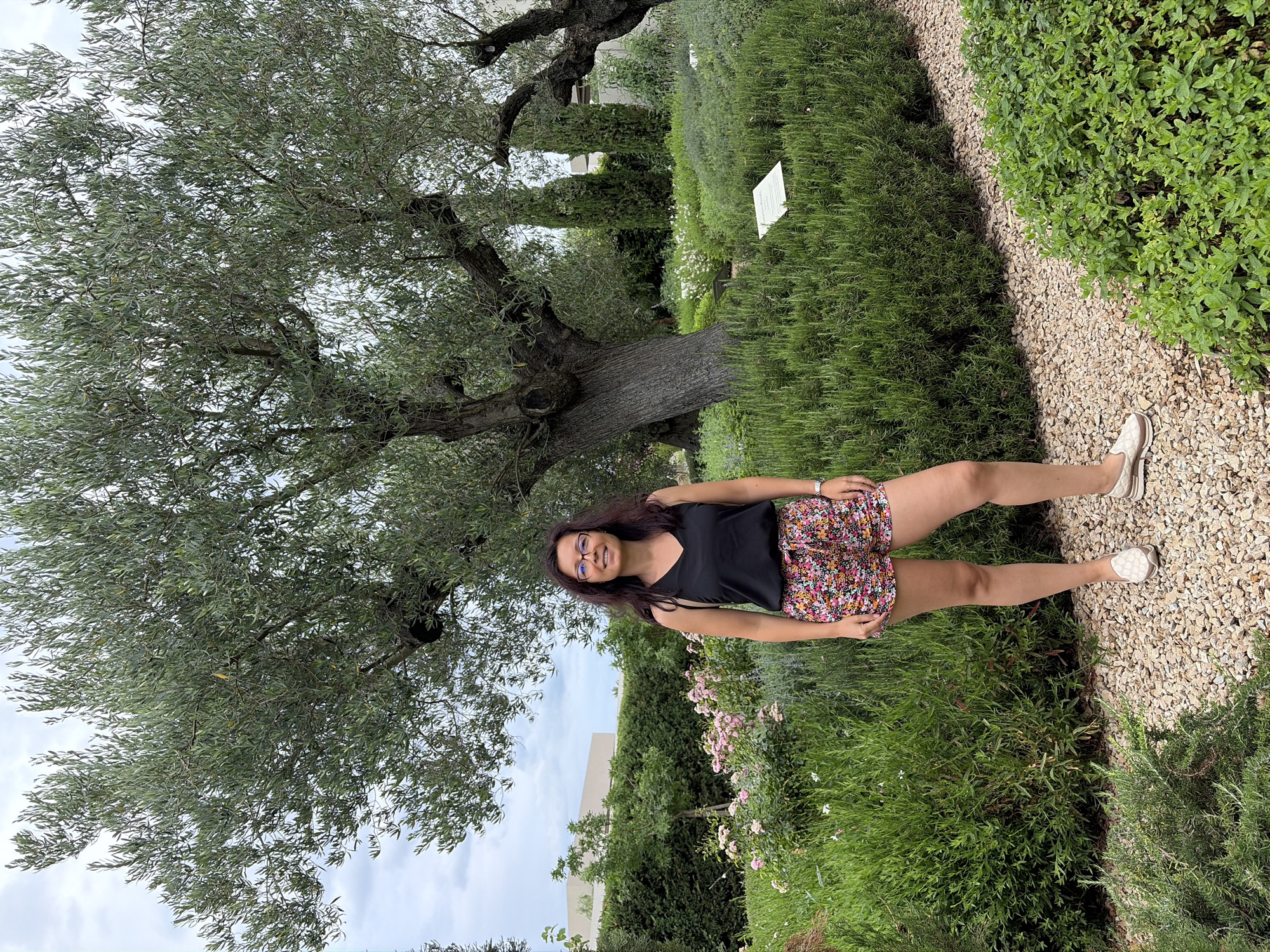 A woman is standing in a garden in front of a large olive tree. She is wearing a black tank top and floral shorts. The garden is filled with various plants and flowers. In the background, there is a building. The sky is cloudy. This scene is located in the city of Kassandra Municipal Unit, Greece.
