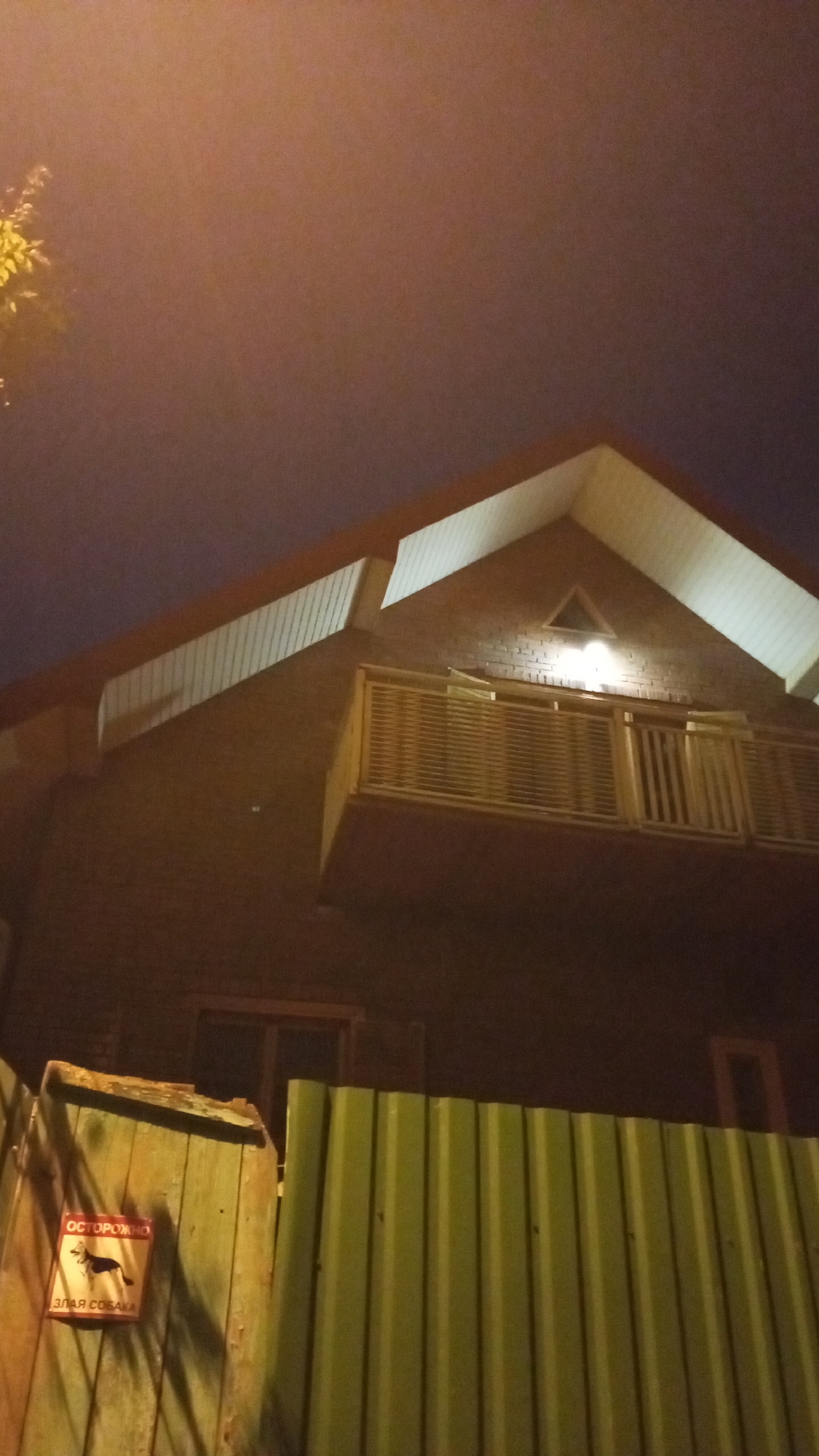 The photograph depicts a building in Юдино, Russia at night. A two-story structure, possibly residential, is visible with a brick exterior and a balcony illuminated by a light positioned above it. Above the balcony is a triangular shape, also illuminated. A fence, composed of corrugated metal, stands in the foreground. To the left of the metal fence is a wooden fence with a sign that reads "ОСТОРОЖНО ЗЛАЯ СОБАКА". A tree branch is visible in the top left corner. The sky is dark and overcast.