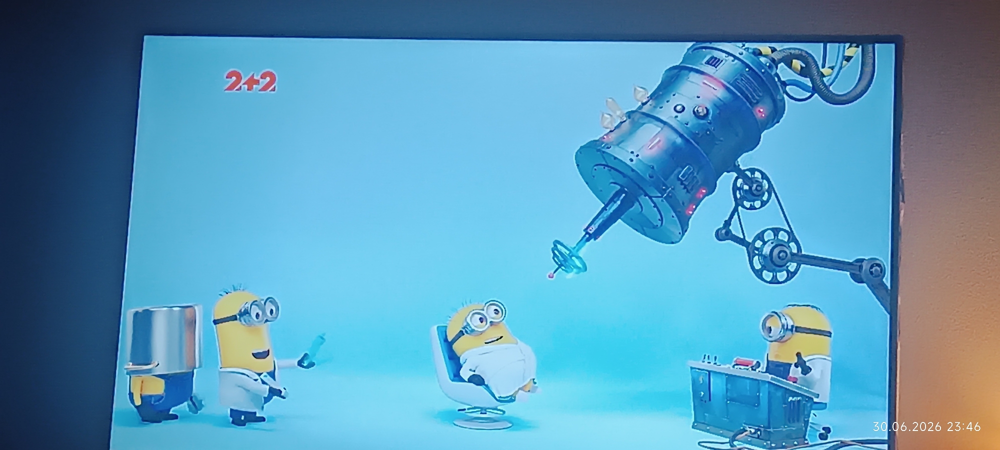 The image depicts a scene featuring several Minions characters from the animated movie franchise. In the upper left corner, the logo "2+2" is visible. The central part of the image is dominated by a large, metallic, cylindrical machine with pipes and gears, resembling a complex apparatus.
In the foreground, three Minions are present. On the left, a Minion wearing a lab coat and goggles holds a syringe. To its right, another Minion, also in a lab coat and goggles, sits in a white chair, appearing to be a patient. Further to the right, a third Minion stands behind a control panel with various buttons and screens, wearing a lab coat and goggles.
The background is a plain, light blue color. The overall scene suggests a laboratory or medical setting, with the Minions engaged in some sort of scientific or medical procedure. There is no discernible urban environment or location in Odesa, Ukraine, as the image is a still from an animated production and does not depict real-world locations. The bottom right corner of the image displays a timestamp: "30.06.2026 23:46".