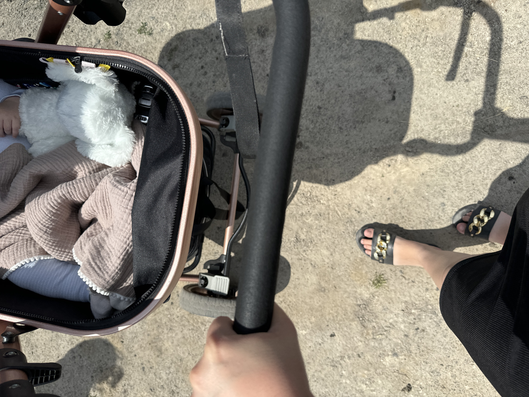 A person's hand is holding the handle of a baby stroller. The stroller contains a baby wrapped in a pink blanket and a white stuffed animal. The person is wearing black sandals with gold chain accents and a black skirt. The stroller is on a concrete path, with shadows cast by the stroller and the person. The scene appears to be outdoors in a city.