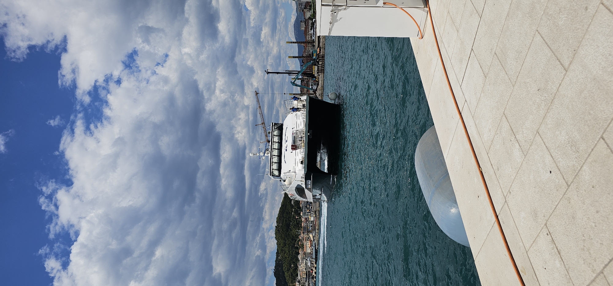 A wide-angle shot captures a large, white catamaran ferry approaching a dock in the city of Split, Croatia. The ferry, with dark hull accents and the text "KATALINA" visible on its side, is moving from left to right across the frame. Two individuals are standing on the deck of the ferry. The water around the ferry is a deep turquoise, with white wakes trailing behind its propulsion.
In the foreground, a light-colored stone-tiled dock extends into the frame, with a large, gray, rounded fender or bumper visible partially submerged in the water. An orange hose or cable runs along the edge of the dock.
The background features a coastal city with buildings nestled on a green, tree-covered hillside. Industrial elements, including a tall yellow crane and construction equipment, are visible on the right side of the frame. The sky is a dramatic mix of bright blue patches and dark, heavy clouds. The overall lighting suggests it is daytime, possibly late morning or early afternoon, with intermittent sunlight.
