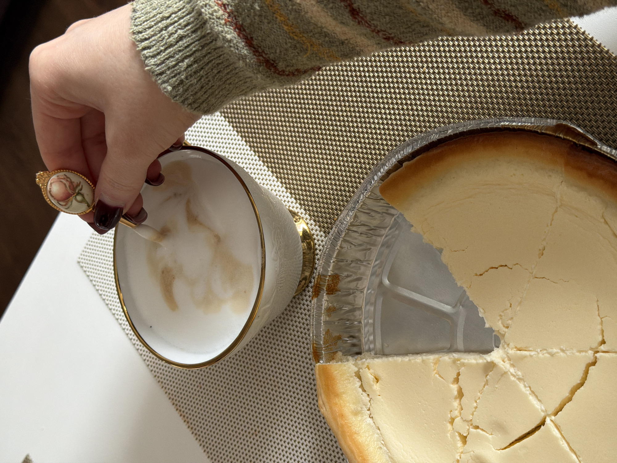 This high-angle shot captures a person's hand stirring a frothy drink, alongside a partially eaten cheesecake, in what appears to be an indoor setting.
The hand, adorned with dark red nail polish, holds an ornate golden spoon featuring a delicate pink rose painting on its handle. The spoon is immersed in a white, gold-rimmed cup, stirring a light-colored beverage with a foamy top, likely a latte or cappuccino. The person is wearing a knitted sweater in shades of olive green, brown, and subtle reddish stripes, visible on the forearm.
To the right, a round cheesecake rests in an aluminum foil pan. A triangular slice has been removed, revealing the creamy, pale yellow interior. The remaining cheesecake is pre-cut into several radiating sections, with some natural cracks visible on its smooth surface. The edges of the cheesecake show a lightly browned, baked crust.
Both the cup and the cheesecake are placed on a gold-colored, woven-textured placemat, which sits on a plain white table surface. The lighting suggests a bright, perhaps sunlit, indoor environment. There is no visible text in the image. The scene depicts a relaxed moment, possibly a coffee or dessert break. The setting is presumably in Penza, Russia.