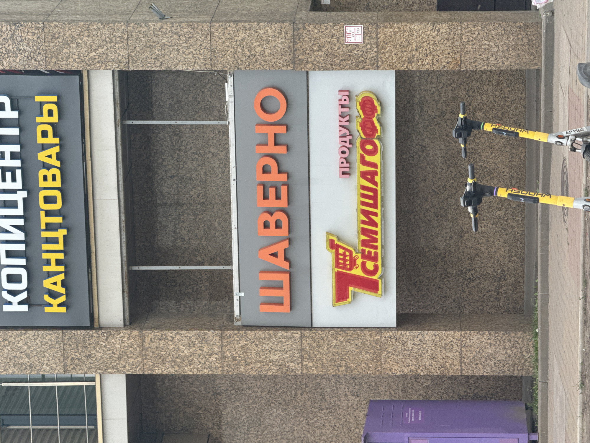 The image displays a daytime street scene in Saint Petersburg, Russia, showing the ground floor of a commercial building with several storefronts. No people are visible.
The scene features three distinct businesses marked by large signs. From left to right (as if the image were rotated upright):
1. A sign with "КОПИЦЕНТР" (Kopitsentr, meaning Copy Center) in white letters and "КАНЦТОВАРЫ" (Kanctovary, meaning Stationery or Office Supplies) in yellow letters on a dark grey background.
2. A sign for "ШАВЕРНО" (Shaverno, meaning Shawarma, a fast-food type) with large orange letters on a grey background.
3. A sign for "ПРОДУКТЫ" (Produkty, meaning Groceries or Products) in pink letters above a large red and yellow logo for "7 СЕМИШАГОФФ" (7 Semishagoff, a chain of discount supermarkets, meaning Seven Steps).
In front of these storefronts, two yellow and black electric scooters are parked on the tiled sidewalk. Each scooter has the letters "НЅООНМ" (HSOOHM) visible on its yellow frame, likely indicating a rental company. The scooter on the right also has a label "AP1160". The sidewalk is paved with light brown tiles, and a small strip of grass with a curb is visible on the far right edge of the image. A purple utility box is partially visible at the bottom. Above the "Шаверно" sign, a small white rectangular sign with "СГ" and numbers "236", "115" is affixed to the building. The lighting suggests an overcast day or indirect sunlight.