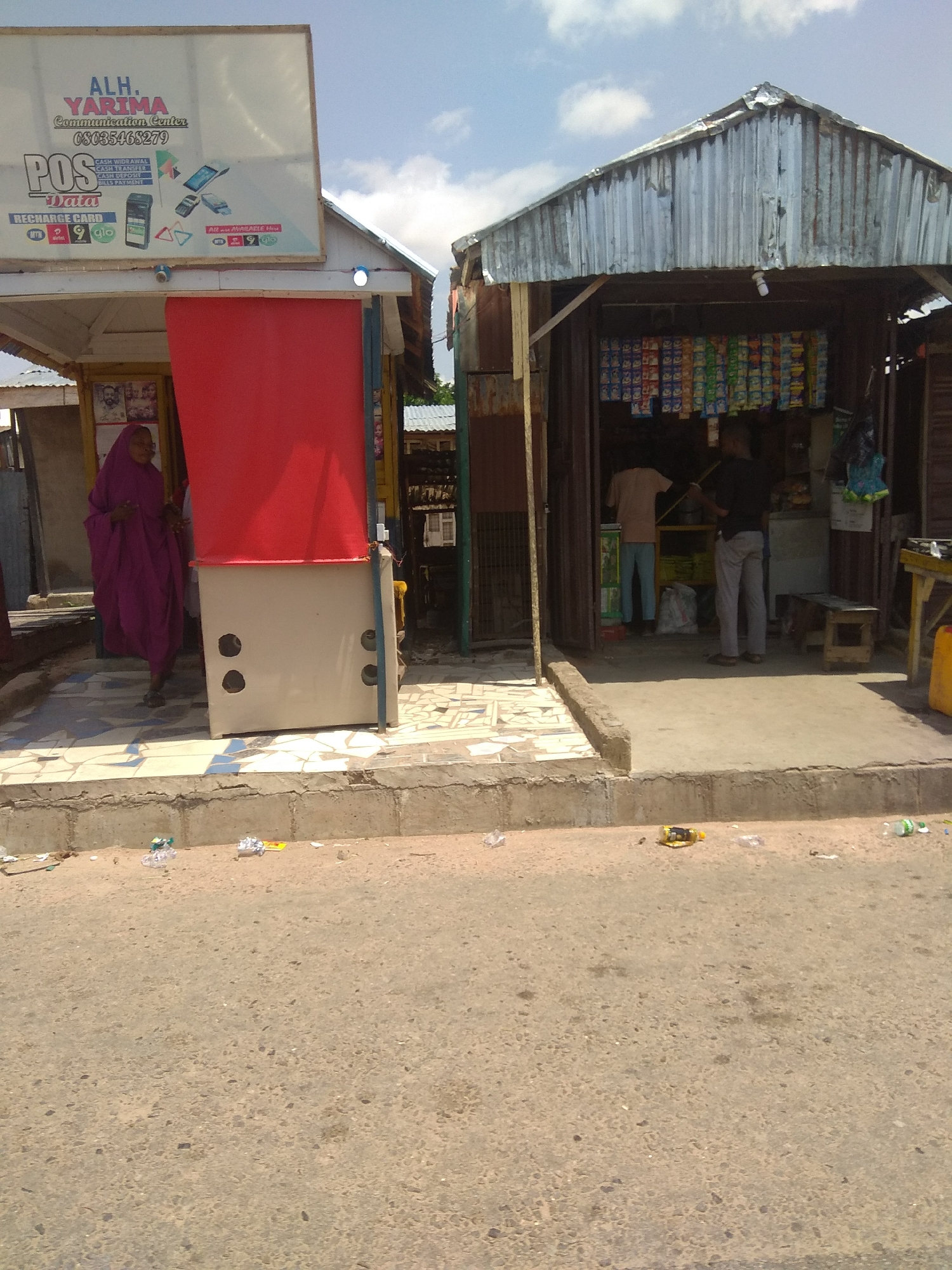 This is a full-body, eye-level, medium shot of an outdoor market stall in Jalingo, Nigeria. The scene is captured on a sunny day. The market stall on the left is a small building with a white sign that reads "ALH. YARIMA COMMUNICATION CENTER". Below the sign are graphics of a POS machine, a mobile phone, and a SIM card, indicating services like cash withdrawal, cash transfer, cash deposit, and airtime payment. A woman wearing a purple hijab and abaya stands in front of a red and white structure, likely part of the stall's display. To the right, another stall features a corrugated metal roof and displays various packaged goods on shelves. Two men are visible inside this stall; one in a light brown shirt and blue pants, and another in a dark shirt and grey pants. The ground in front of the stalls is paved with tiles, and the street in the foreground is asphalted. Litter is scattered along the edge of the street.