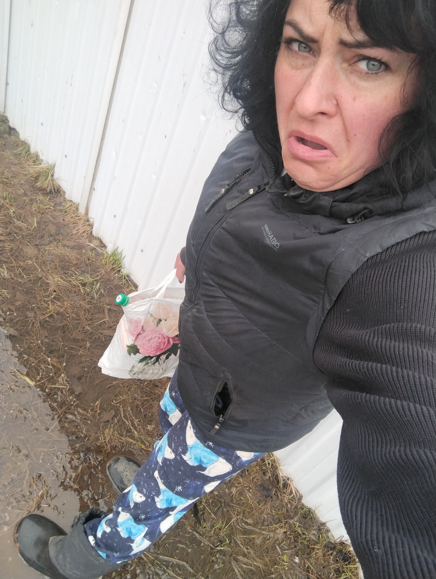 A woman is standing in a muddy area near a white metal fence. She is wearing black rubber boots, blue and white patterned pants, a black sweater, and a black vest. She is holding a white plastic bag with pink flowers and a green bottle inside. Her facial expression indicates disgust. The background consists of mud, grass, and a white metal fence. The location is Grishanki, Russia.