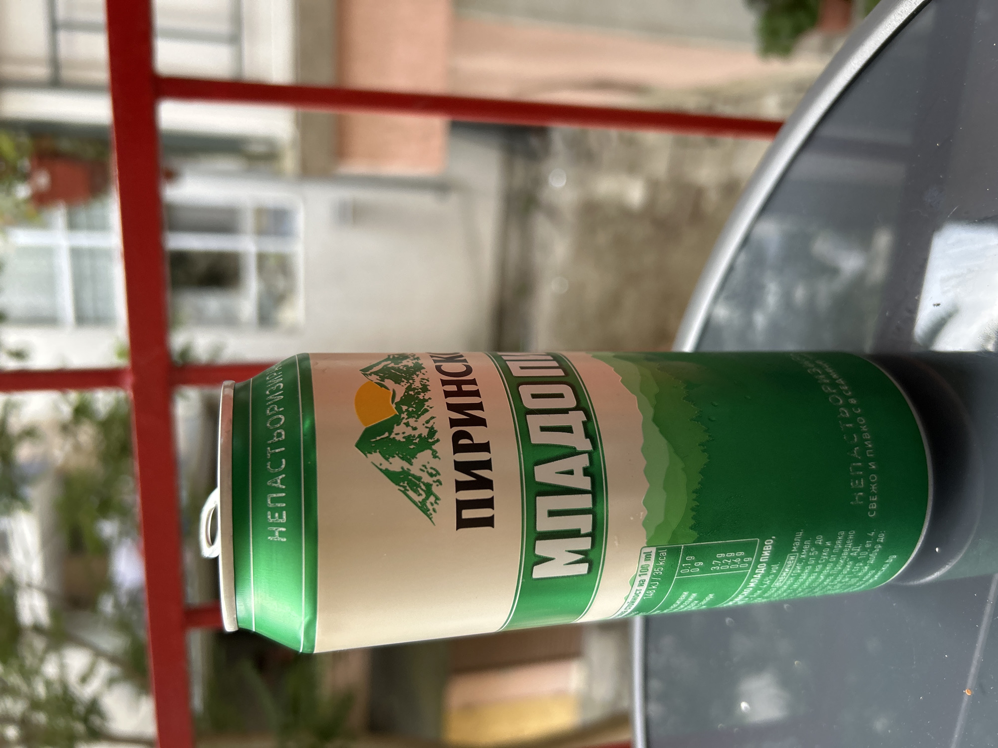 A metallic green and cream-colored can of Pirinsko Mlado Pivo is positioned on a dark, reflective table outdoors. The can features a mountain landscape graphic with a sun setting behind the peaks. Text in Cyrillic is visible on the can, including "НЕПАСТЬОРИЗИРАНО" and "ПИРИНСКО МЛАДО ПИВО". A nutritional information table is also present on the lower portion of the can, listing values per 100 ml. The background shows a blurred urban environment with a red railing, buildings, and greenery, possibly in the city of Burgas, Bulgaria, as indicated by the beer brand's origin. The lighting suggests it is daytime.