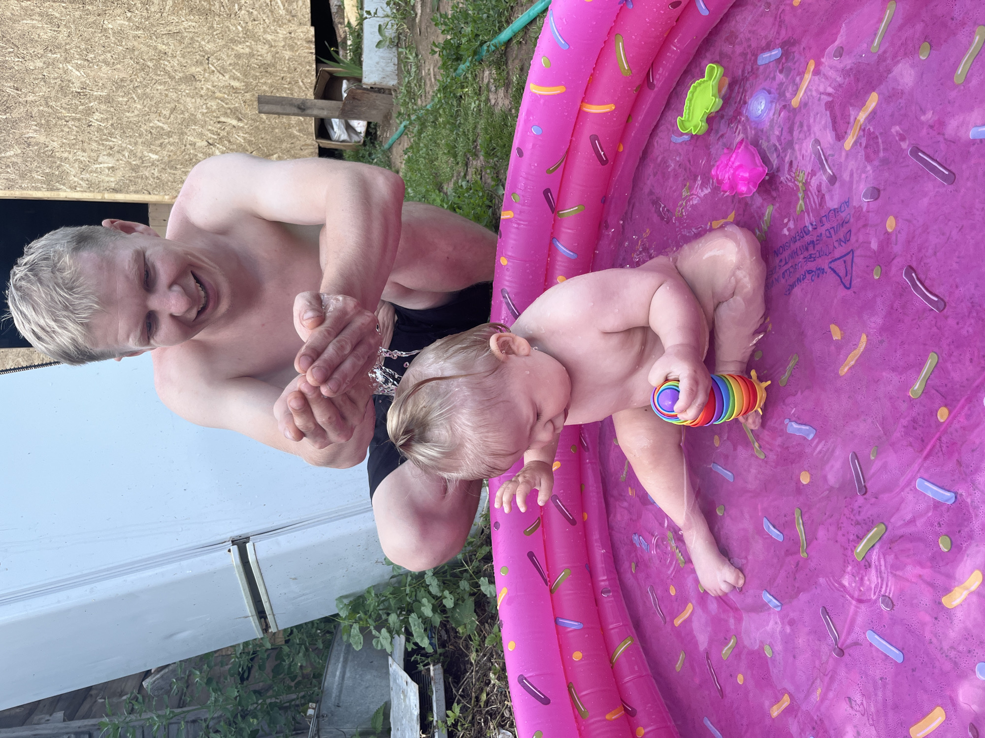 In Фоки, Russia, a man with blond hair, shirtless, crouches over an inflatable pink kiddie pool. His hands are cupped, holding water over a baby. The baby, also shirtless, sits in the pool, holding a rainbow-colored toy. The pool is decorated with sprinkle-like patterns. The background shows an outdoor environment with a white refrigerator, wooden structures, and greenery. The scene appears to be a backyard on a warm day.