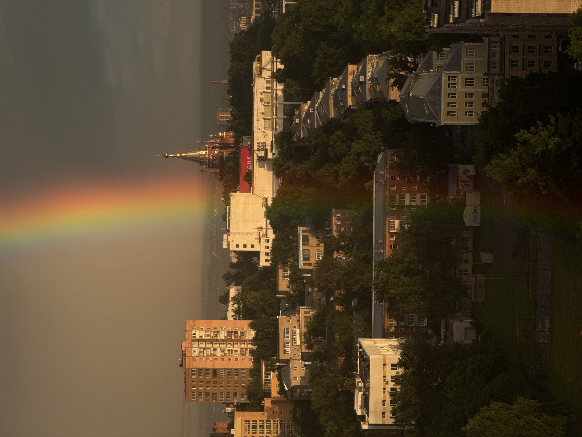 The aerial view captures a cityscape in Izhevsk, Russia, dominated by a vibrant rainbow arcing across a dusky sky. The rainbow's spectrum of colors, from red to violet, is prominently displayed, cutting diagonally through the frame. Below, a dense collection of urban architecture unfolds. Various apartment buildings, characterized by brick and concrete facades, are interspersed with lush green trees. One prominent building, a brick apartment complex with numerous windows, stands out in the left mid-ground. Further in the background, a Russian Orthodox church with golden domes gleams under the dramatic lighting. The city stretches out towards the horizon, veiled in a soft haze. The scene appears to be captured during or just after a rain shower, as indicated by the rainbow and the overcast sky. No people are visible in the frame.