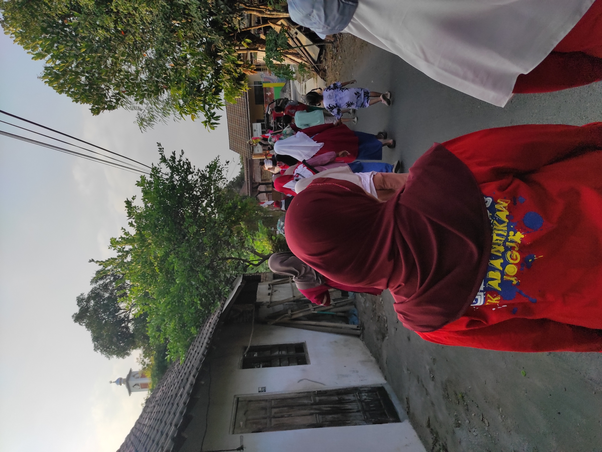 The image shows a group of people, mostly women and children, walking down a paved street in what appears to be a village. The architecture suggests a location in Southeast Asia, possibly Indonesia, with one building having a tiled roof and wooden doors. In the background, there's a tall, slender tower with a dome and a clock, resembling a minaret, suggesting a mosque nearby. The overall lighting indicates it's either early morning or late afternoon, with the sun casting a soft glow. The people are dressed in casual clothing, with many of the women wearing hijabs. The scene depicts a leisurely walk or a community gathering.
