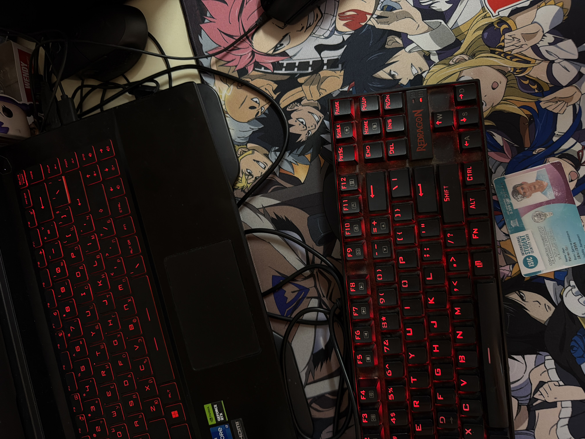The image shows a close-up, overhead view of a gaming setup. A black laptop with a red-backlit keyboard is positioned above a black gaming keyboard with red backlighting. The gaming keyboard is placed on top of a mousepad featuring characters from an anime series. To the right of the laptop, a black computer mouse is visible. On the mousepad, slightly to the right of the center, is a partially visible ISIC International Student Identity Card, with the name "Ivan" and a photo of a young man. The background is dark, suggesting an indoor setting. The lighting is dim, with the primary illumination coming from the red backlighting of the keyboards and possibly a faint overhead light. There are no discernible city landmarks or specific details that would place the scene in Sofia, Bulgaria. The overall scene depicts a personal gaming or computer workspace.