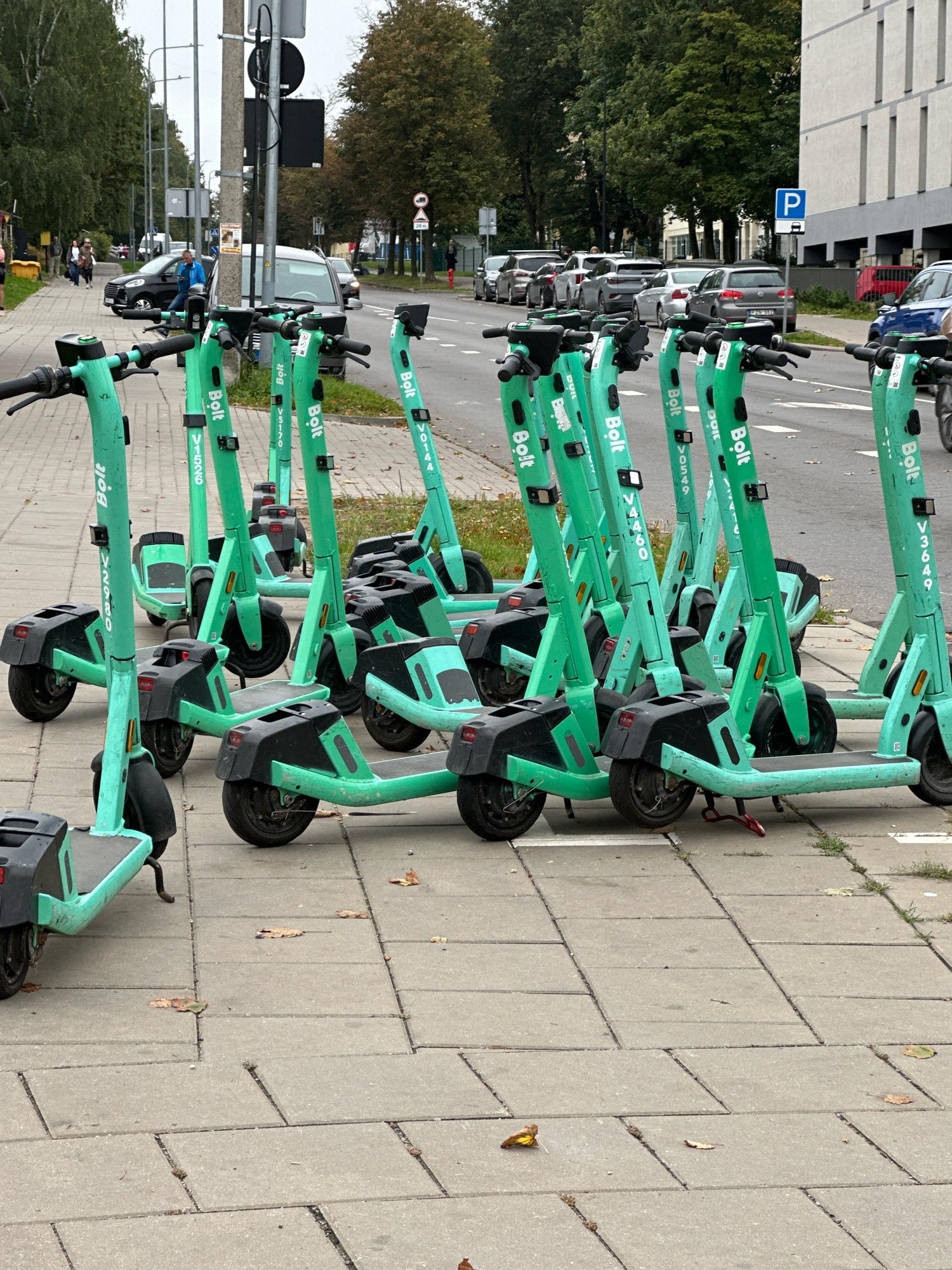 The image shows a row of green electric scooters from the company Bolt parked on a sidewalk in Vilnius, Lithuania. The scooters are lined up neatly, with their handlebars facing forward. Several of the scooters have visible identification numbers on their frames, such as "V1526", "V0549", and "V0144". In the background, a street is visible with cars parked along the side and trees lining the sidewalk. A building with a modern facade is also present on the right side of the image, and a parking sign with a blue 'P' is displayed. A traffic light pole with a black traffic signal is visible on the left side of the frame. The sky is overcast, suggesting a cloudy day. Some fallen leaves are scattered on the sidewalk, indicating it might be autumn. There are no people actively interacting with the scooters in the foreground.
