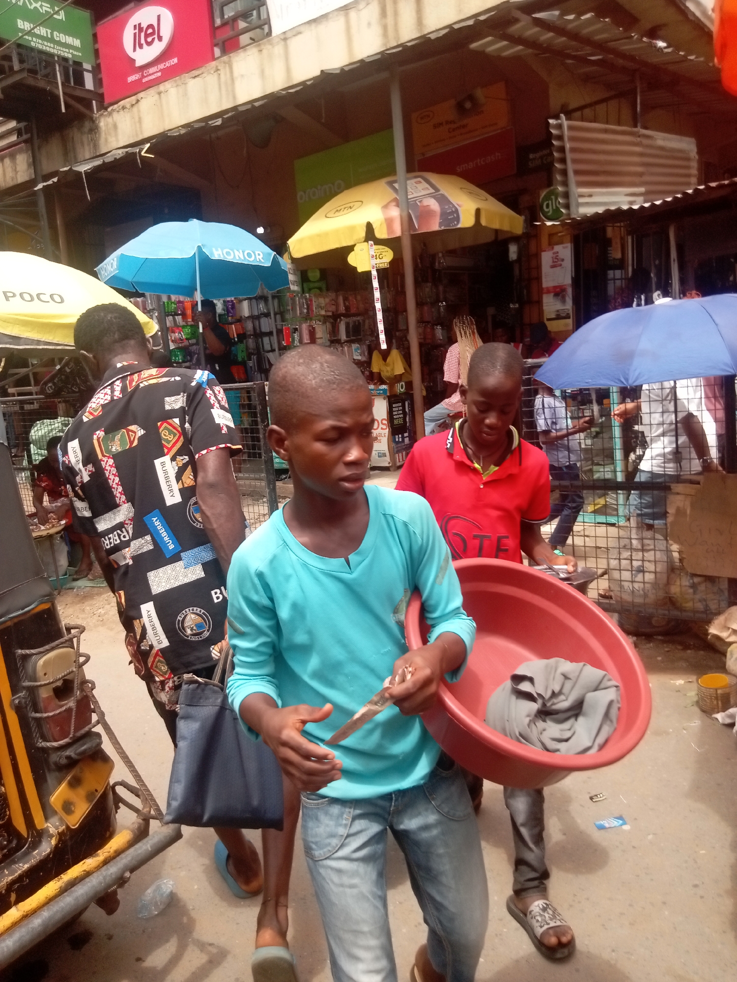 This image depicts a bustling street market scene in Fegge, Nigeria, likely during the daytime. The bright lighting and numerous umbrellas (blue "HONOR," yellow "POCO," and yellow "MTN") suggest sunny weather and the need for shade.
In the foreground, a young boy wearing a light blue long-sleeve shirt and jeans holds some small items and a large red plastic basin containing a gray cloth. Beside him, another young boy in a red polo shirt with "TE" visible on it is looking down at something in his hands. A third individual, seen from behind, wears a black short-sleeved shirt patterned with "BURBERRY" logos and carries a dark blue bag. Part of a yellow and black tricycle is visible on the far left.
The background reveals various market stalls or shops, displaying a multitude of phone accessories, such as cases and chargers. Overhead, several signs advertise telecommunication and electronics brands, including a prominent red "itel" sign, a green "BRIGHT COMM" sign with contact details ("400 879/880 Einson Plaza," "07030067598"), a yellow "oraimo" sign, and signs for "MTN," "smartcash," "Register SIM Card," "SIM Registration Center," and "glo." Corrugated metal roofs and cluttered displays add to the vibrant market atmosphere. The people in the image appear focused on their activities, contributing to the lively commercial environment.