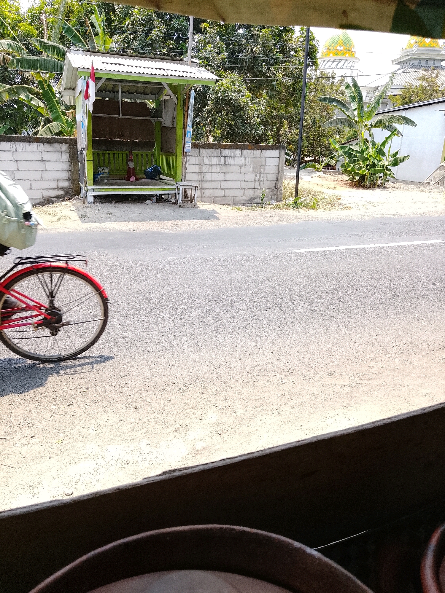 In the city of Banaran, Indonesia, a red bicycle with a rack is in motion on a paved road. The bicycle's rear wheel is blurred due to its movement. To the left of the road, there is a small green shelter with a white roof and a white brick wall behind it. The Indonesian flag, red and white, is displayed on the shelter. Beyond the shelter, trees and the distinctive domes of a mosque can be seen in the background. The foreground of the image shows a brown, curved object, possibly a large pot or container. The scene is illuminated by bright sunlight.