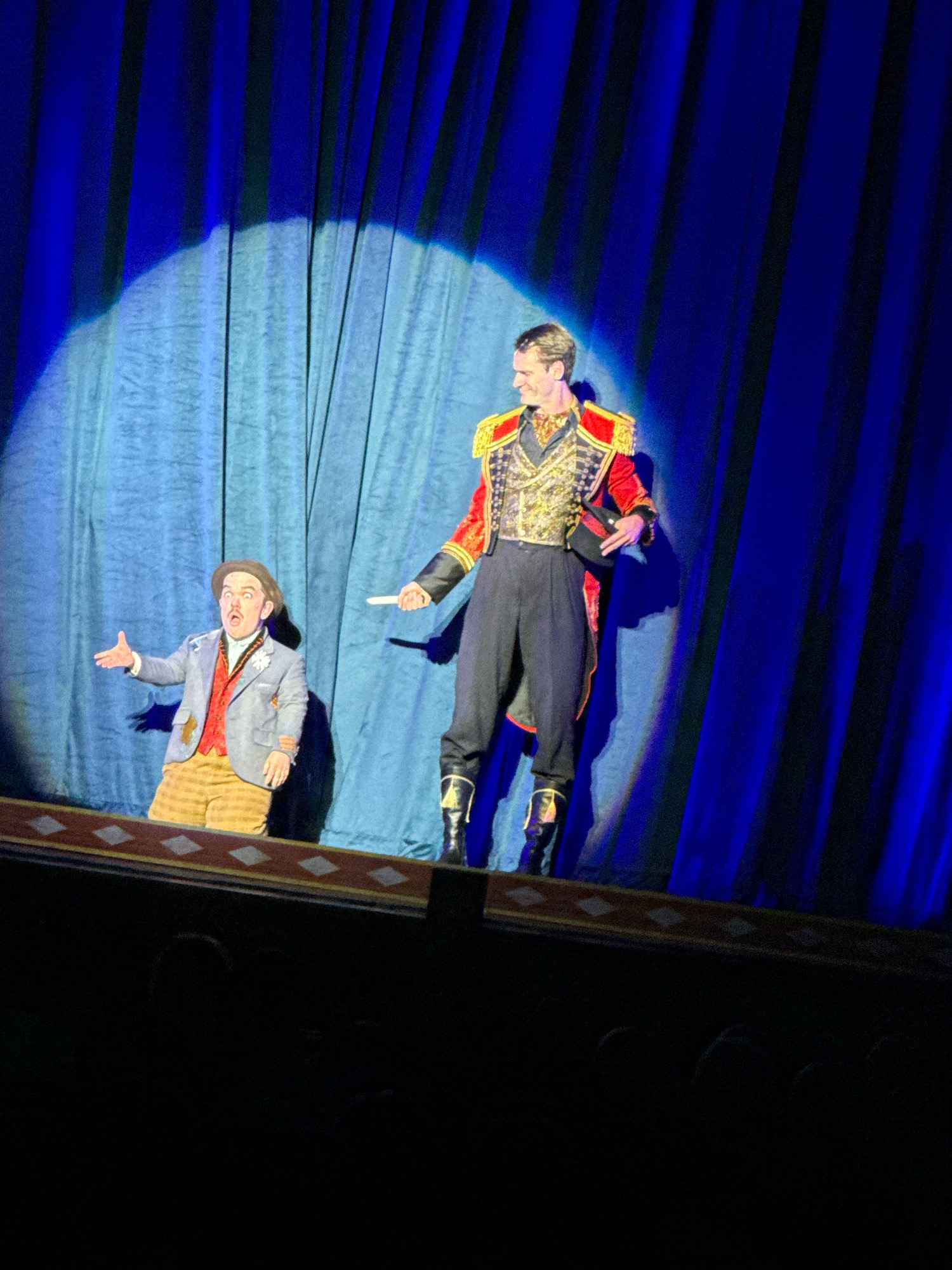 Two performers are on a stage in front of a blue curtain. The performer on the left is a dwarf wearing a grey blazer, a red vest, yellow plaid pants, and a hat. He has a surprised expression and his left arm is outstretched. The performer on the right is a tall man dressed in a red and gold military-style jacket, dark pants, and tall boots. He is looking down and to the left, holding a small white object in his right hand. A spotlight shines on the performers from above. The stage has a decorative border. The image was taken in Los Angeles, California, USA.