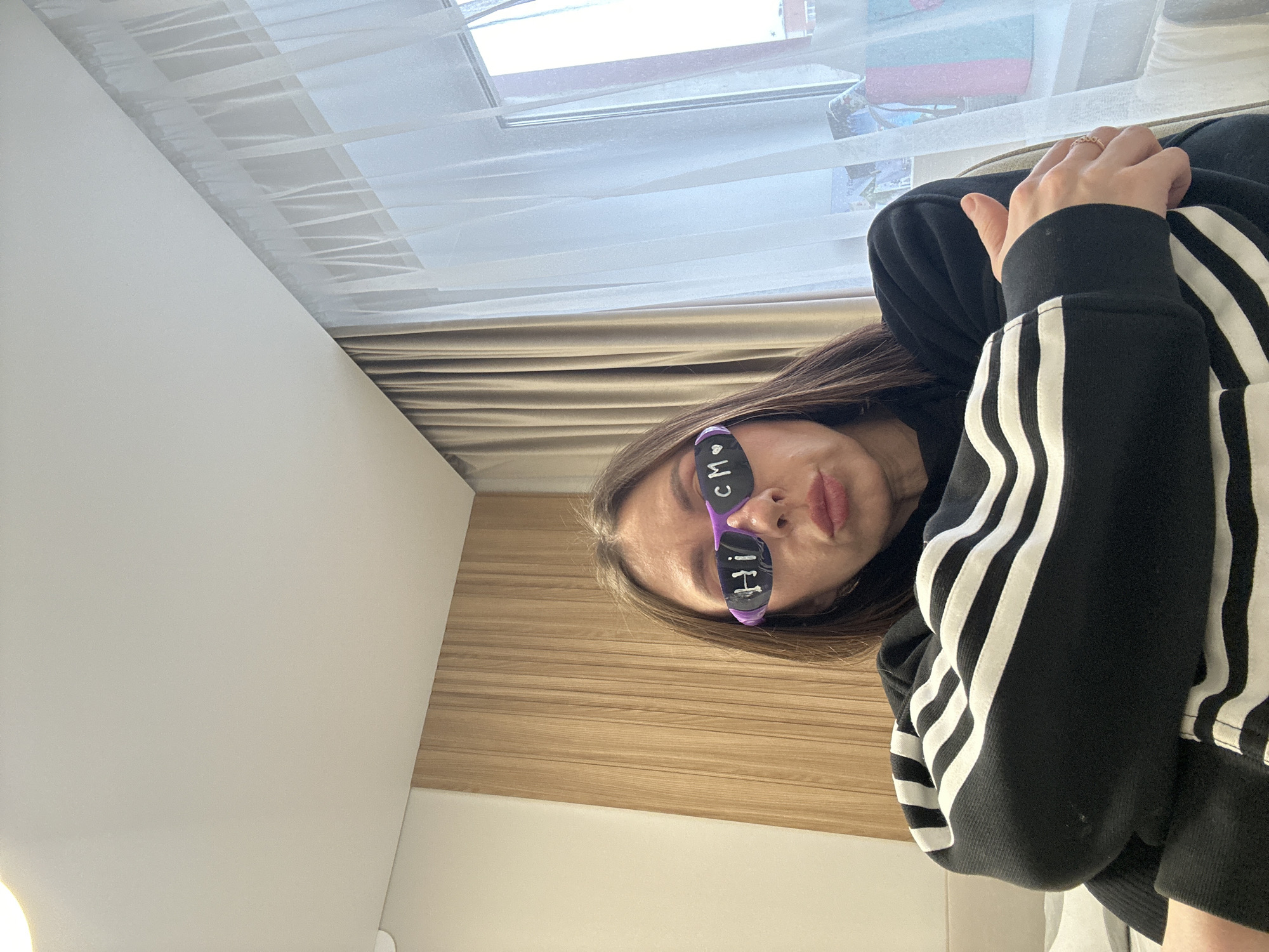 A woman with long brown hair, wearing a black Adidas hoodie with white stripes on the sleeves and purple sunglasses with the words "hi" and "cm" written on them, is angled in the frame. She has a ring on her right hand. The background consists of a window with sheer curtains, wooden wall paneling, and a white wall. The image does not provide sufficient information to determine a specific location within Votkinsk, Russia.