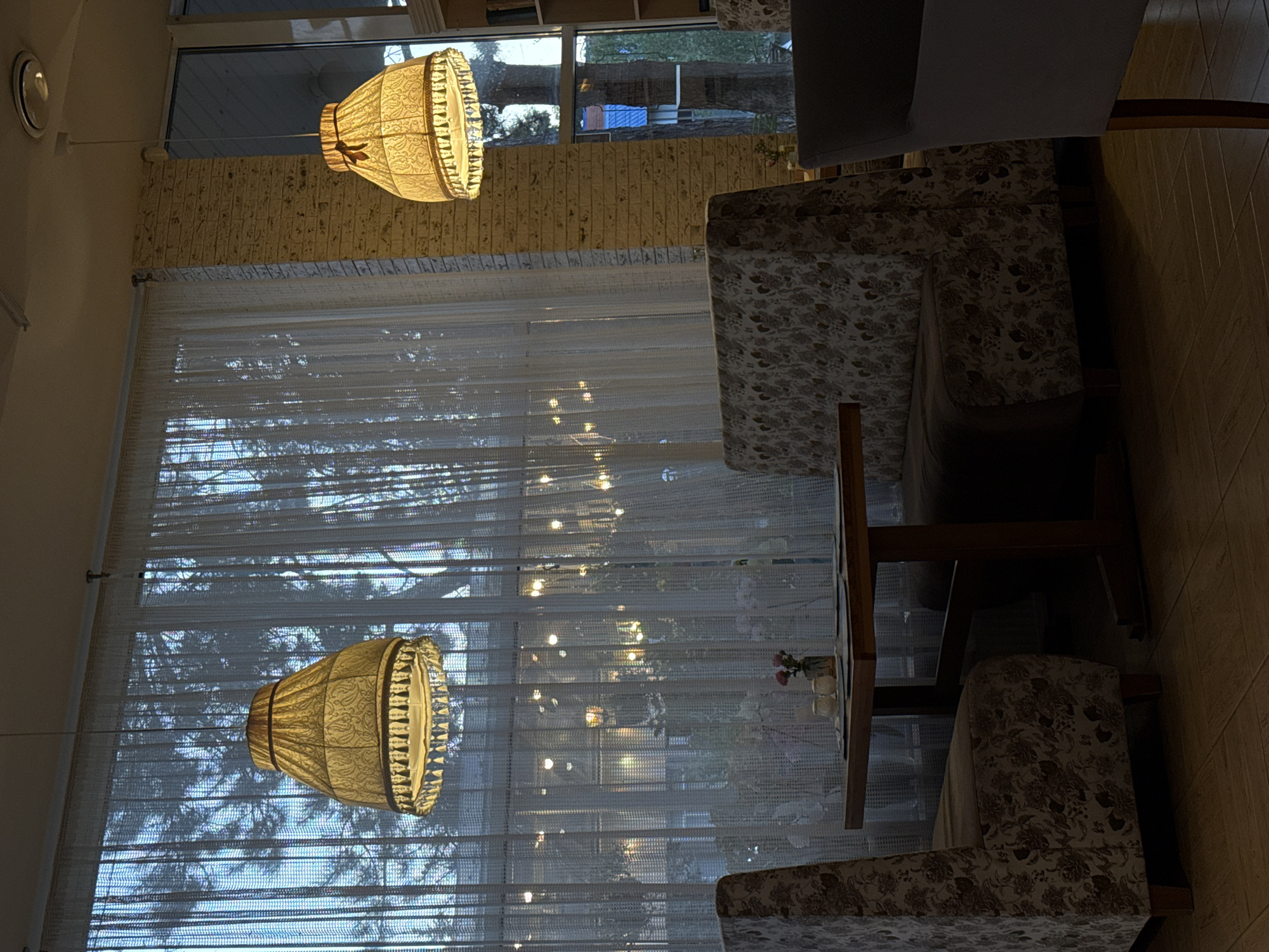The image shows the interior of a cafe in Abinsk, Russia. Two ornate lamps hang from the ceiling, casting a warm glow on the room. In the foreground, a table is set with two salt and pepper shakers and a small vase with pink flowers. Two upholstered chairs with a floral pattern flank the table. Behind the table, a large window is covered by sheer white curtains, through which the silhouettes of trees and outdoor lights can be seen. A brick wall runs along the right side of the room, and a bookshelf is visible in the background. The overall atmosphere is cozy and inviting, suggesting it is evening or a dimly lit day.