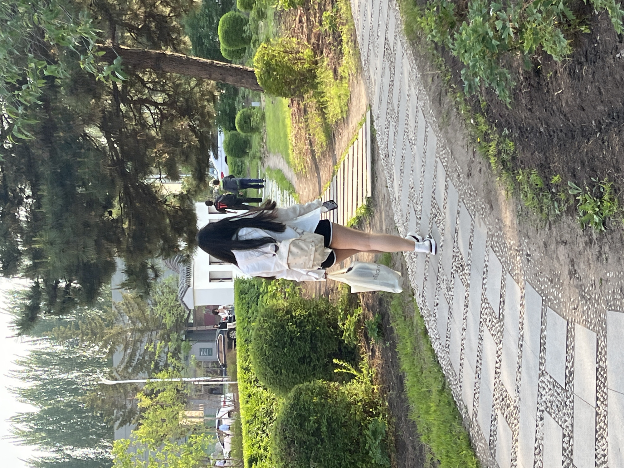 A woman with long dark hair is walking along a stone pathway in Changchun City, China. She is wearing a white shirt, black shorts, white socks, and black and white shoes. She is carrying a light-colored bag over her shoulder and a white plastic bag in her hand. The pathway is lined with green bushes and trees. In the background, there are buildings, cars, and other people.