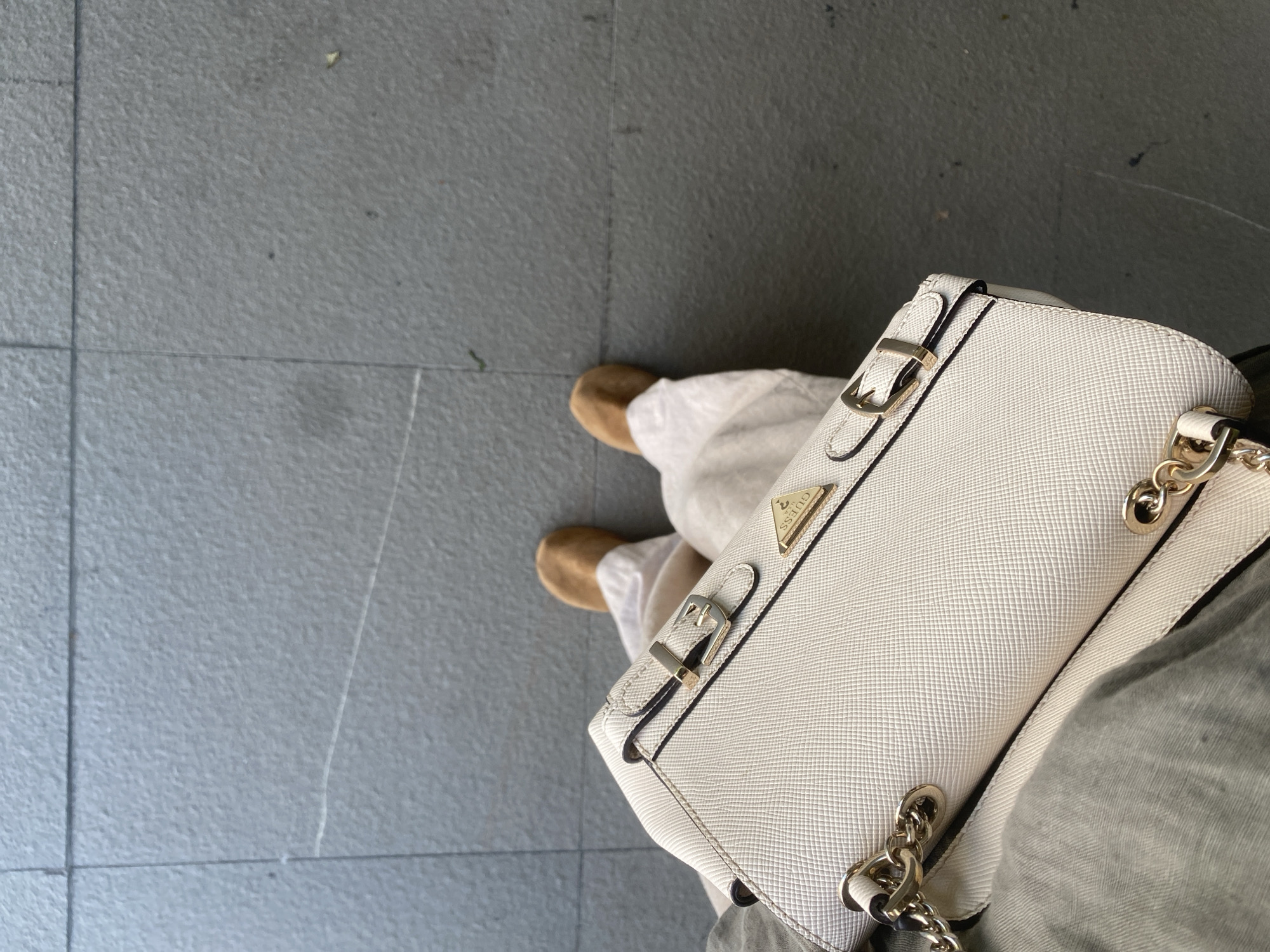 This is a first-person perspective image of a person standing on a tiled ground. The person is wearing light-colored pants and tan shoes. They are holding a white handbag with gold hardware. The handbag has a textured surface and a triangular logo with the word "GUESS" on it. The background is a grey tiled surface. The lighting suggests it is daytime, possibly outdoors. The overall impression is casual and urban.